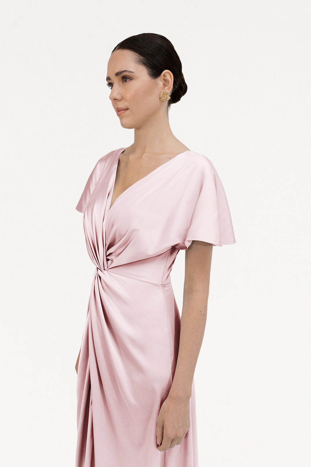 A woman with dark hair in a low bun wears the TH&TH Camilla Bridesmaid Dress in blush pink satin, featuring short sleeves and a twist detail at the waist. She stands against a plain white background, looking slightly to the side.