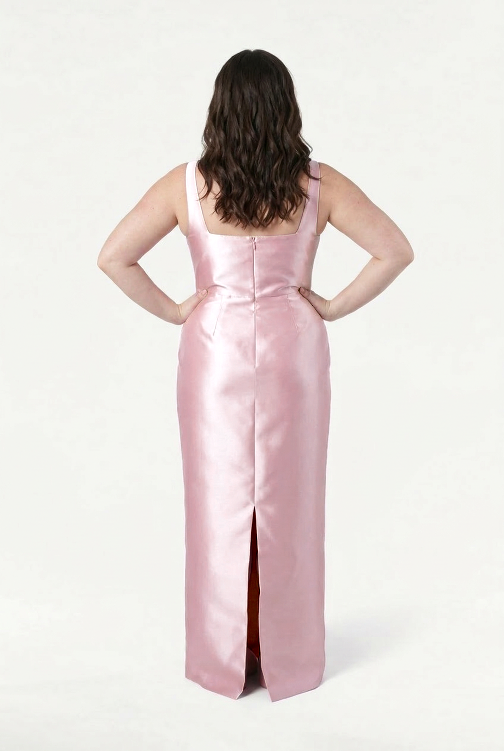 A person with wavy brown hair faces away, hands on hips, wearing the TH&TH Savoy Bridesmaid Dress in Blush Pink Satin Twill featuring a modern square neckline and back slit, set against a plain white background.