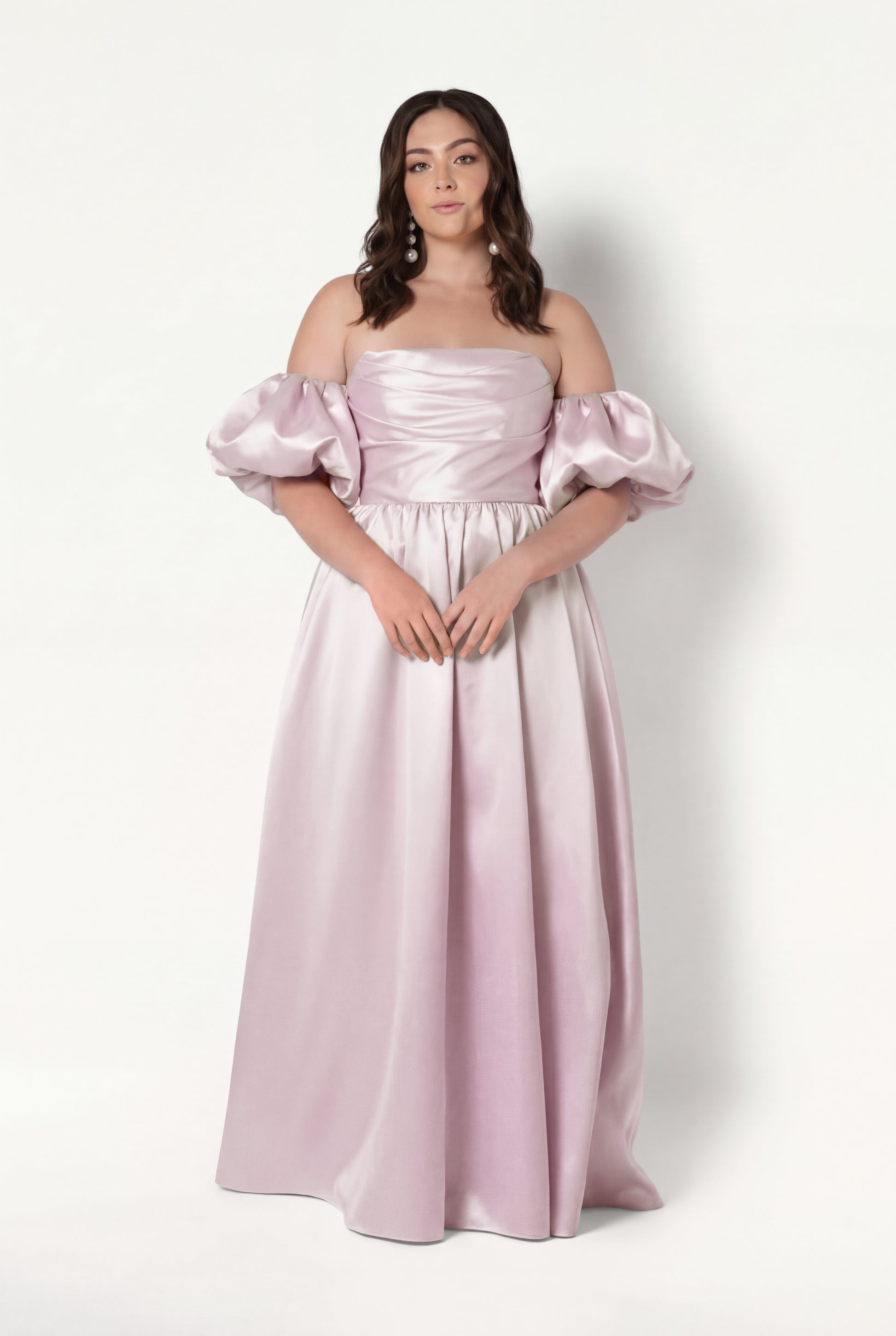 A woman stands against a white background wearing the TH&TH Blair Bridesmaid Dress in Blush Pink Satin Twill, an off-the-shoulder, floor-length gown with detachable sleeves. Her hair is styled in loose waves and her hands rest lightly in front of her.