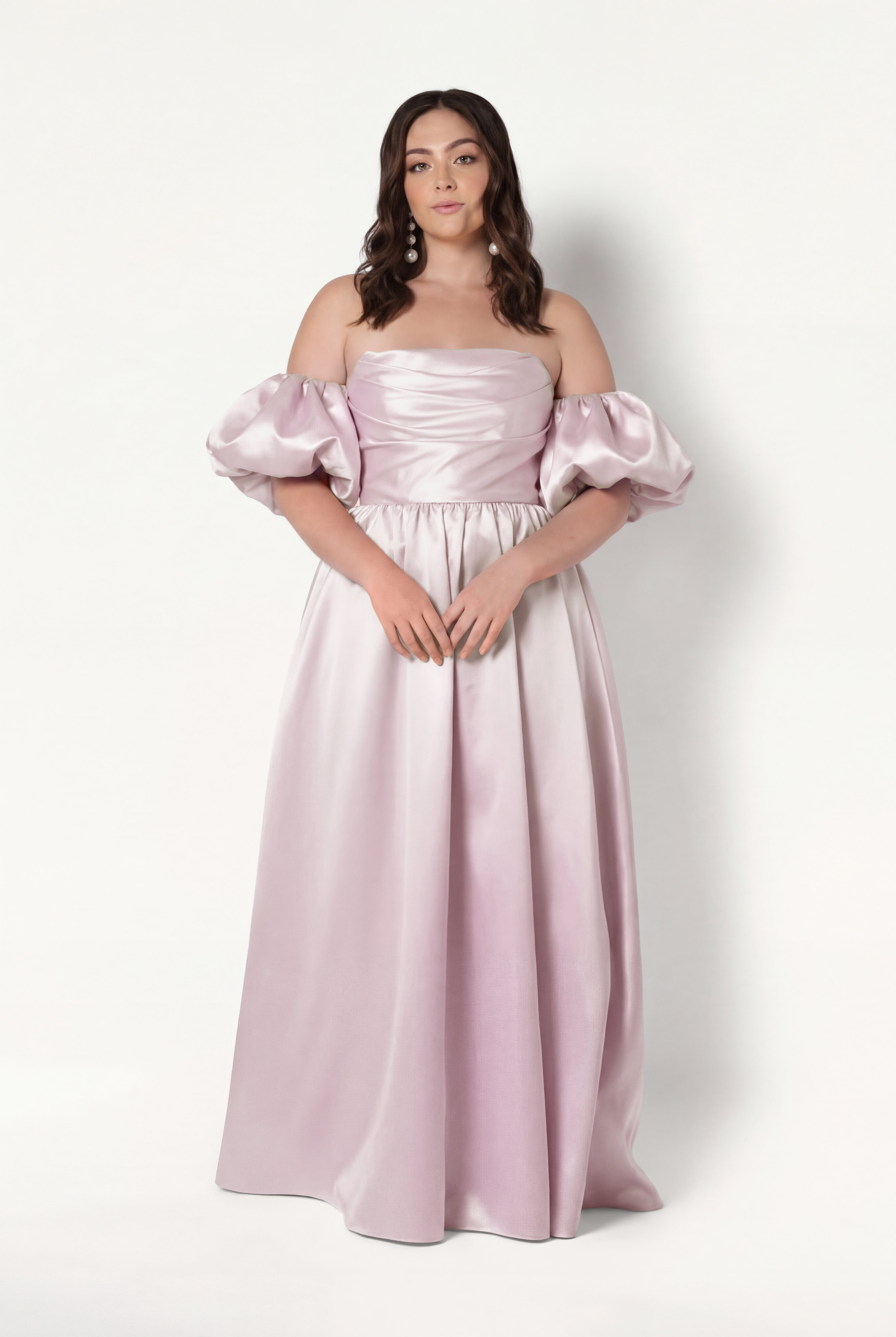 A woman stands against a white background wearing the TH&TH Blair Bridesmaid Dress in Blush Pink Satin Twill, an off-the-shoulder, floor-length gown with detachable sleeves. Her hair is styled in loose waves and her hands rest lightly in front of her.