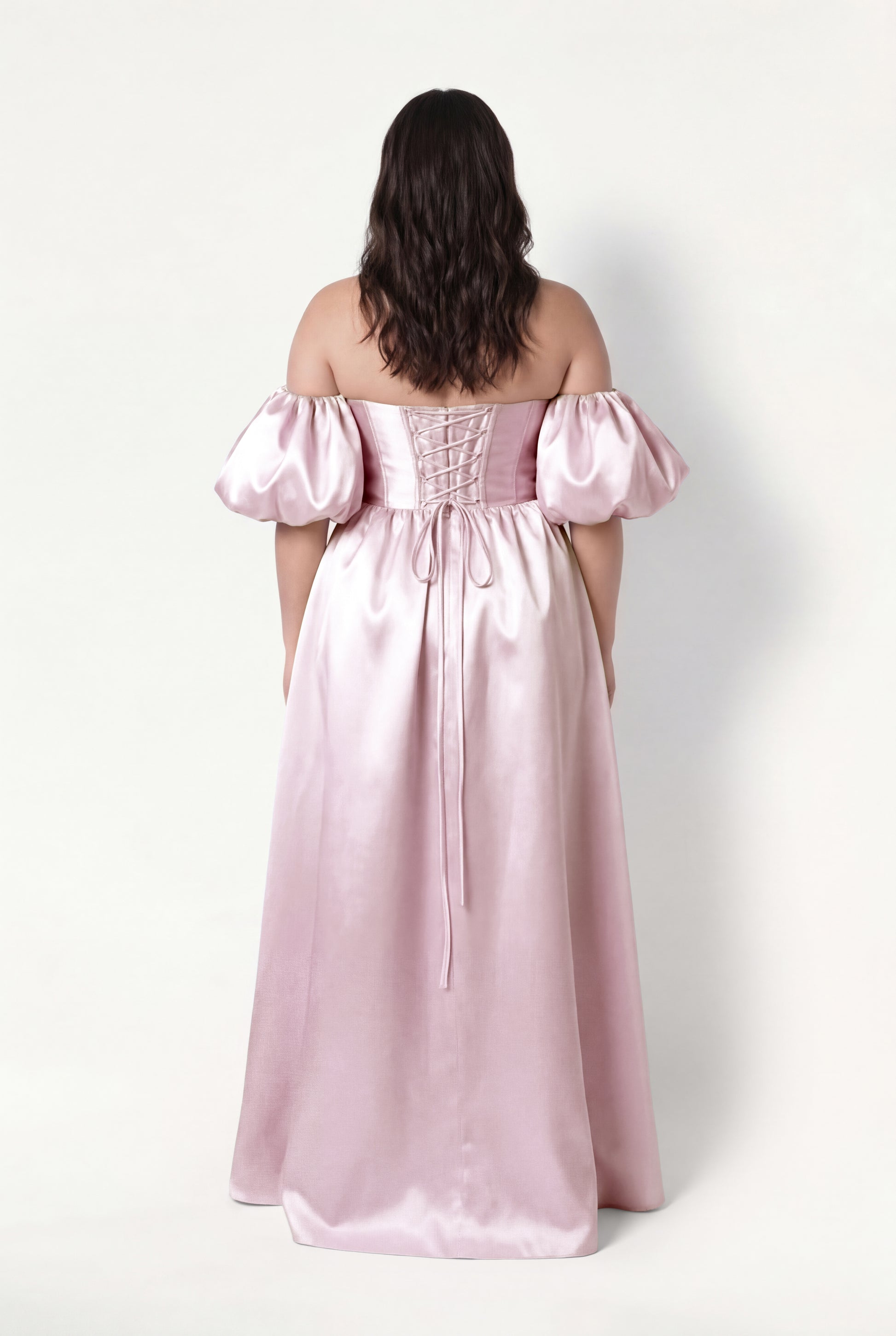 A woman with wavy brown hair faces away, wearing the TH&TH Blair Bridesmaid Dress in blush pink satin twill. The off-the-shoulder gown features detachable puffed sleeves, a lace-up back, and a floor-length, graceful silhouette.
