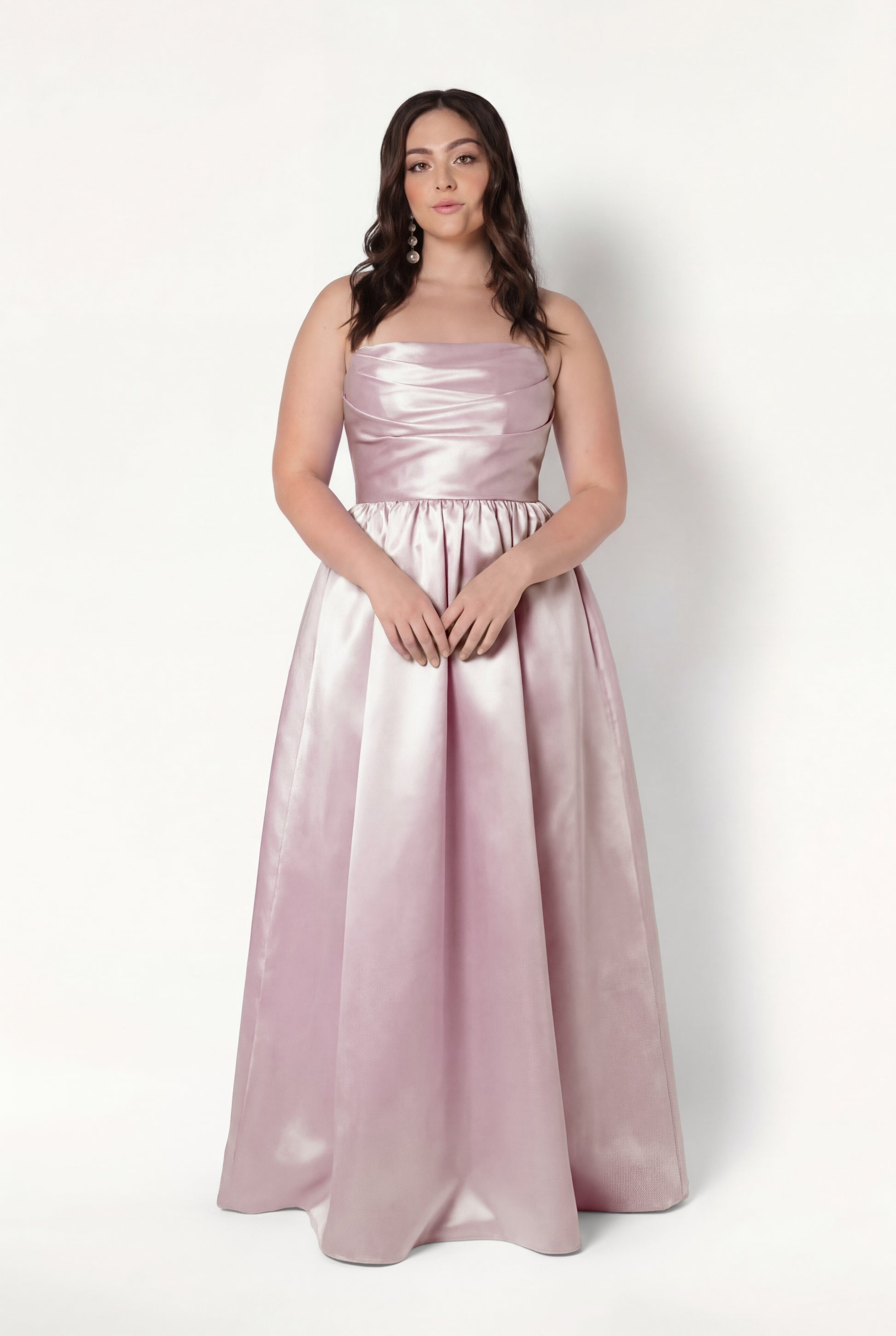 A woman with long, wavy brown hair and a neutral expression wears the TH&TH Blair Bridesmaid Dress in blush pink satin twill, featuring a fitted bodice and full-length skirt, standing against a white background.