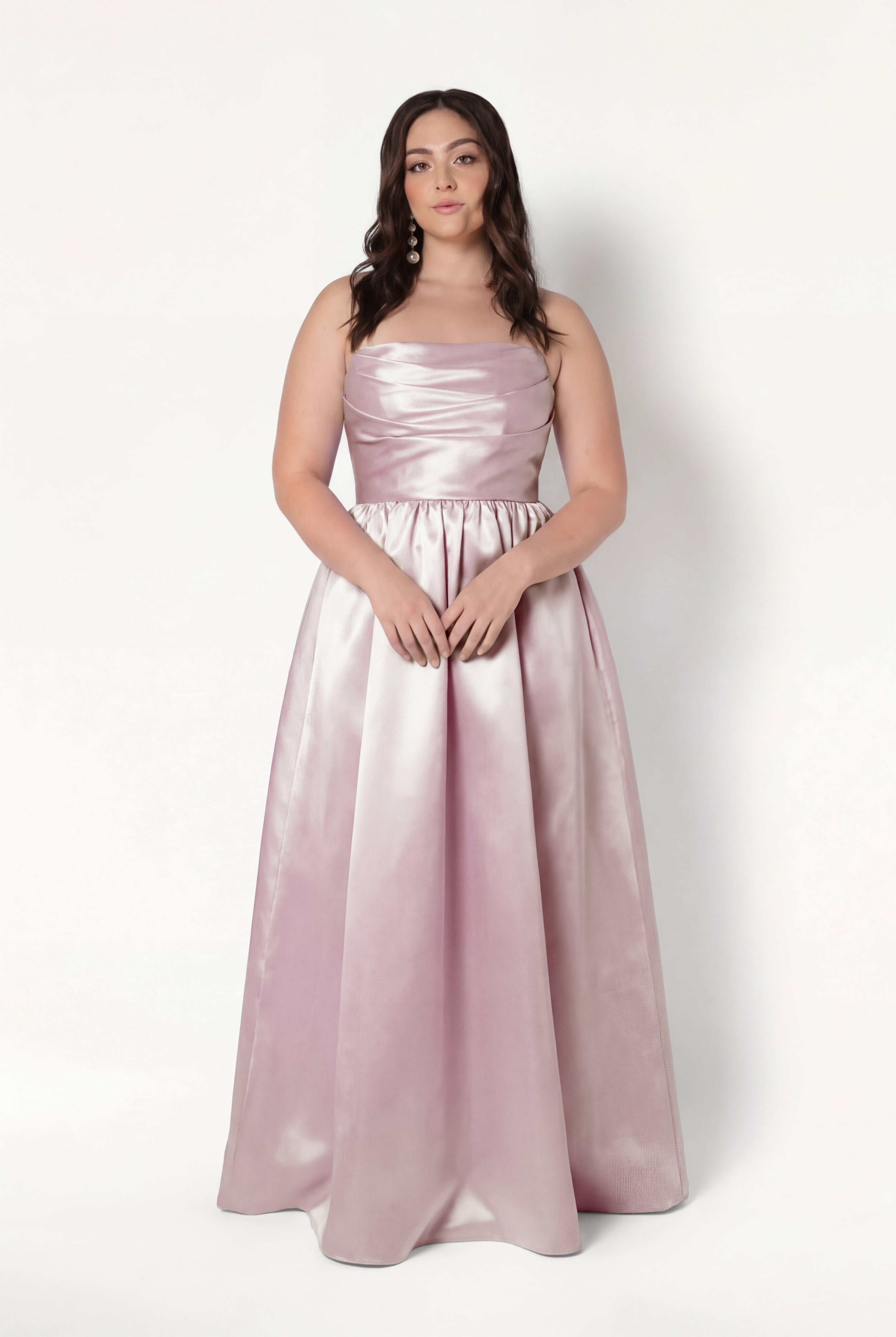 A woman with long, wavy brown hair and a neutral expression wears the TH&TH Blair Bridesmaid Dress in blush pink satin twill, featuring a fitted bodice and full-length skirt, standing against a white background.