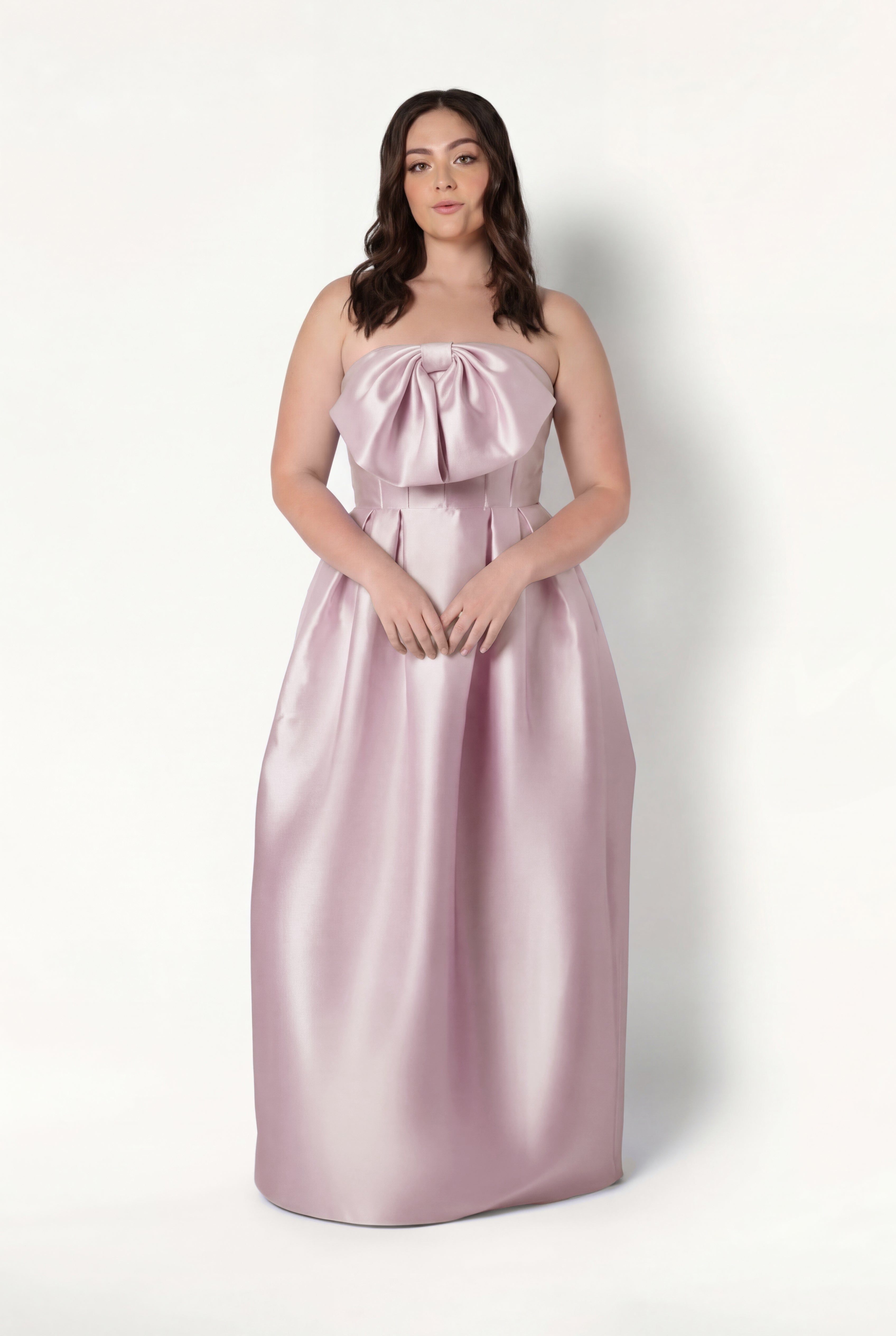 A woman with wavy brown hair wears the TH&TH Ophelia Bridesmaid Dress in Blush Pink Satin Twill, featuring a strapless design and large bow on the bodice, while posing confidently against a plain white background.