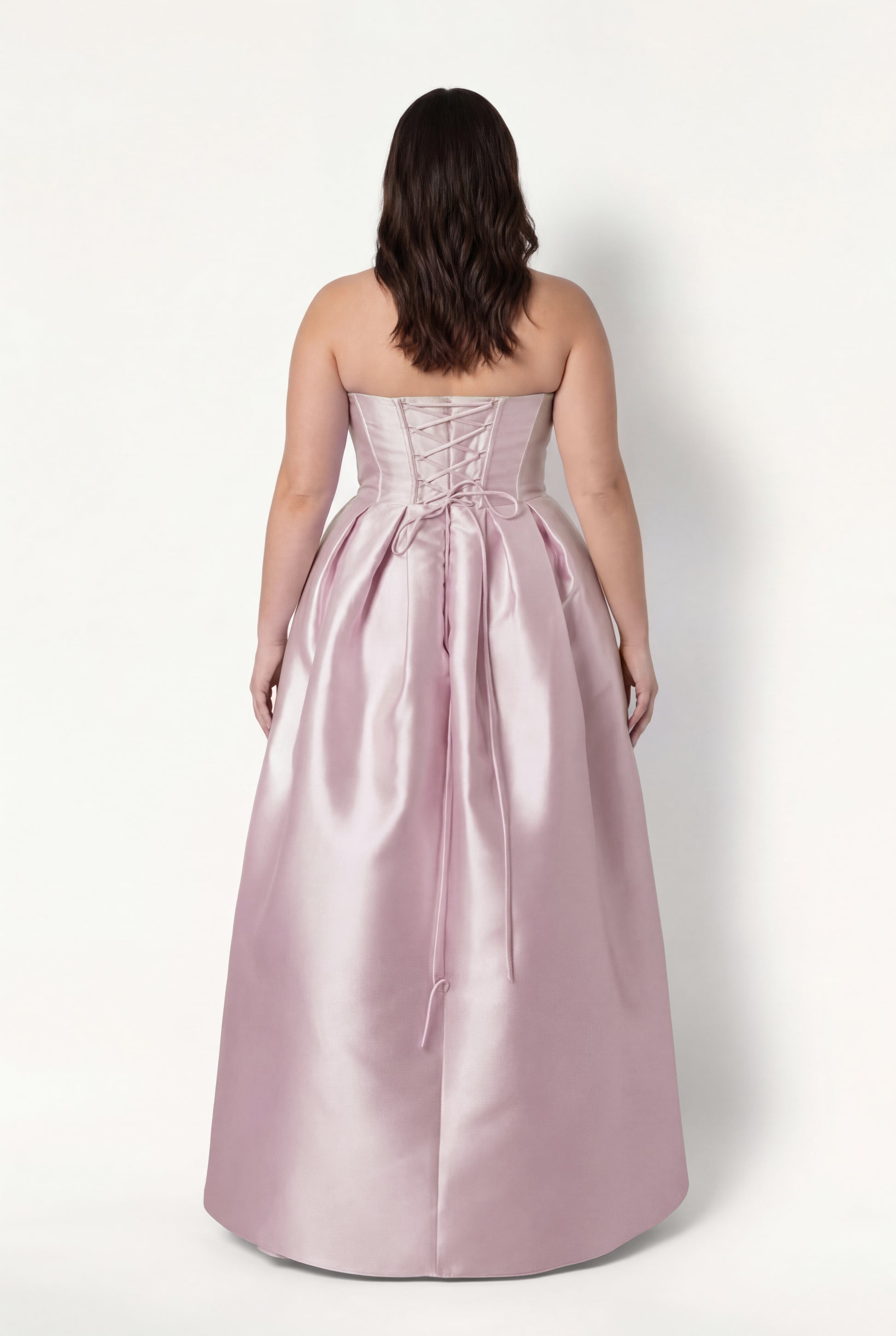 A woman with wavy brown hair models the TH&TH Ophelia Bridesmaid Dress in Blush Pink Satin Twill, featuring a strapless lace-up corset bodice and gathered floor-length skirt, standing against a plain white background, facing away.