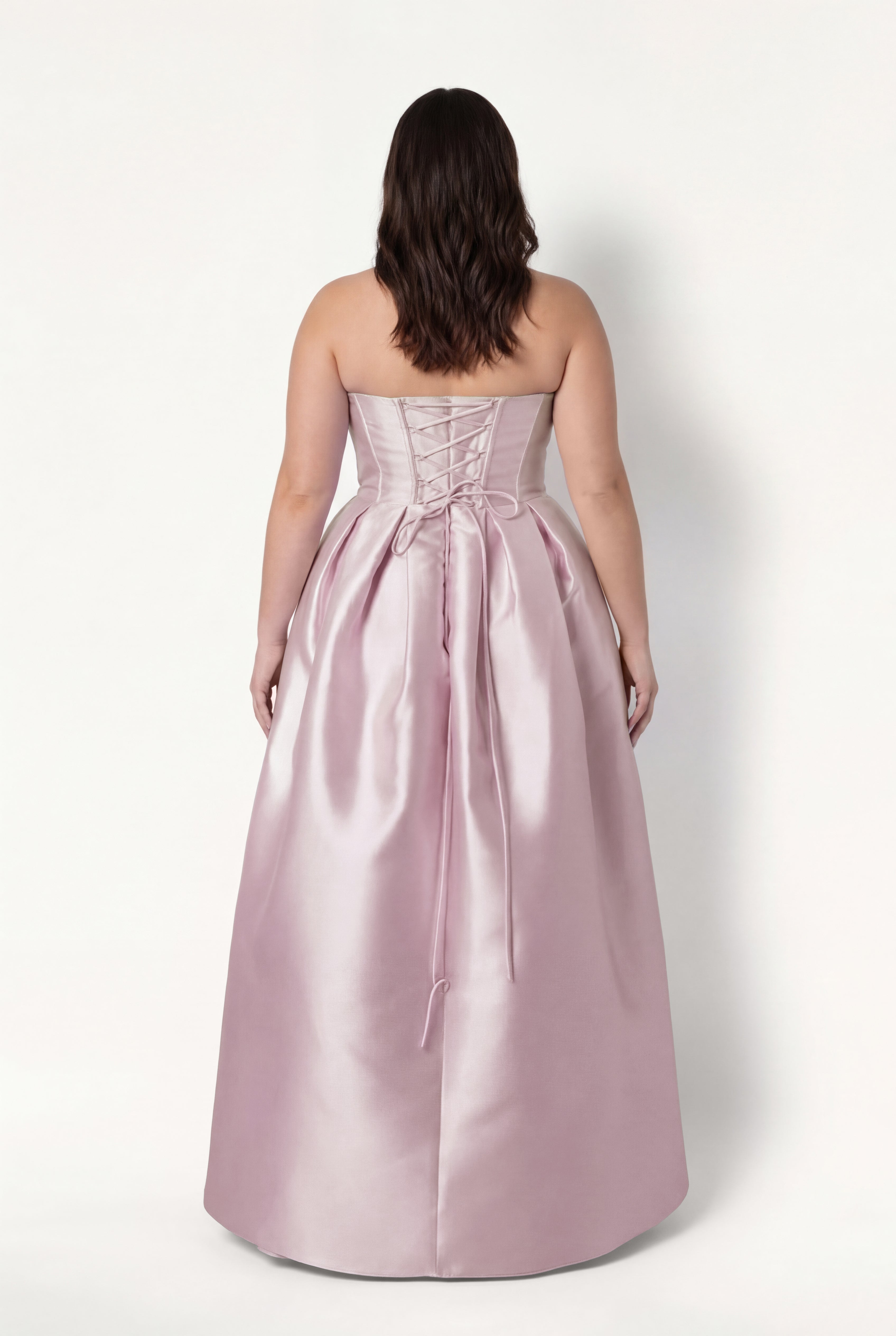 A woman with wavy brown hair models the TH&TH Ophelia Bridesmaid Dress in Blush Pink Satin Twill, featuring a strapless lace-up corset bodice and gathered floor-length skirt, standing against a plain white background, facing away.