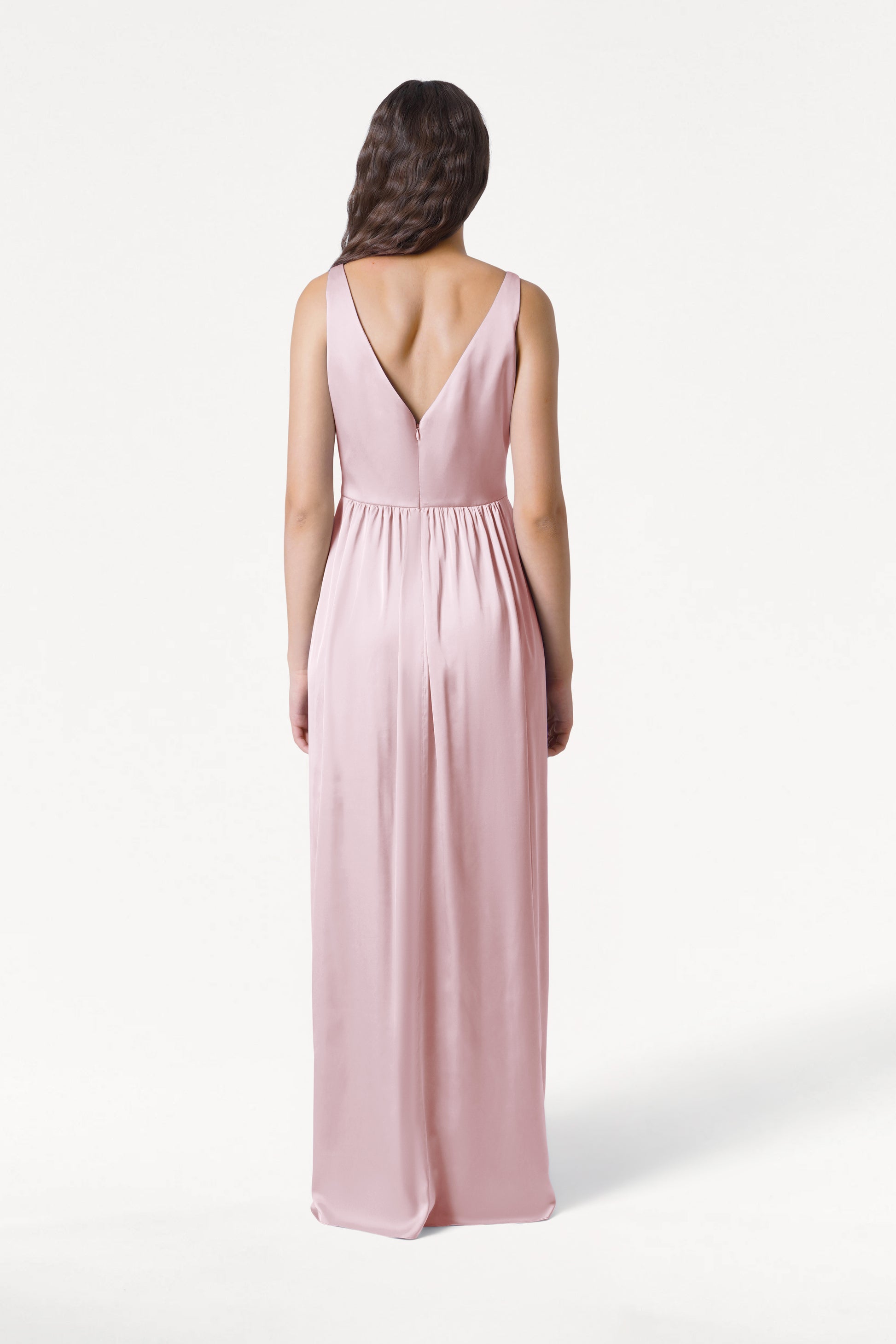 A woman faces away from the camera, wearing the Carmen Bridesmaid Dress in Blush Pink Satin by TH&TH—a long, sleeveless gown with a deep V-back and gathered waist—her loose waves complementing the modern bridal look against a white background.