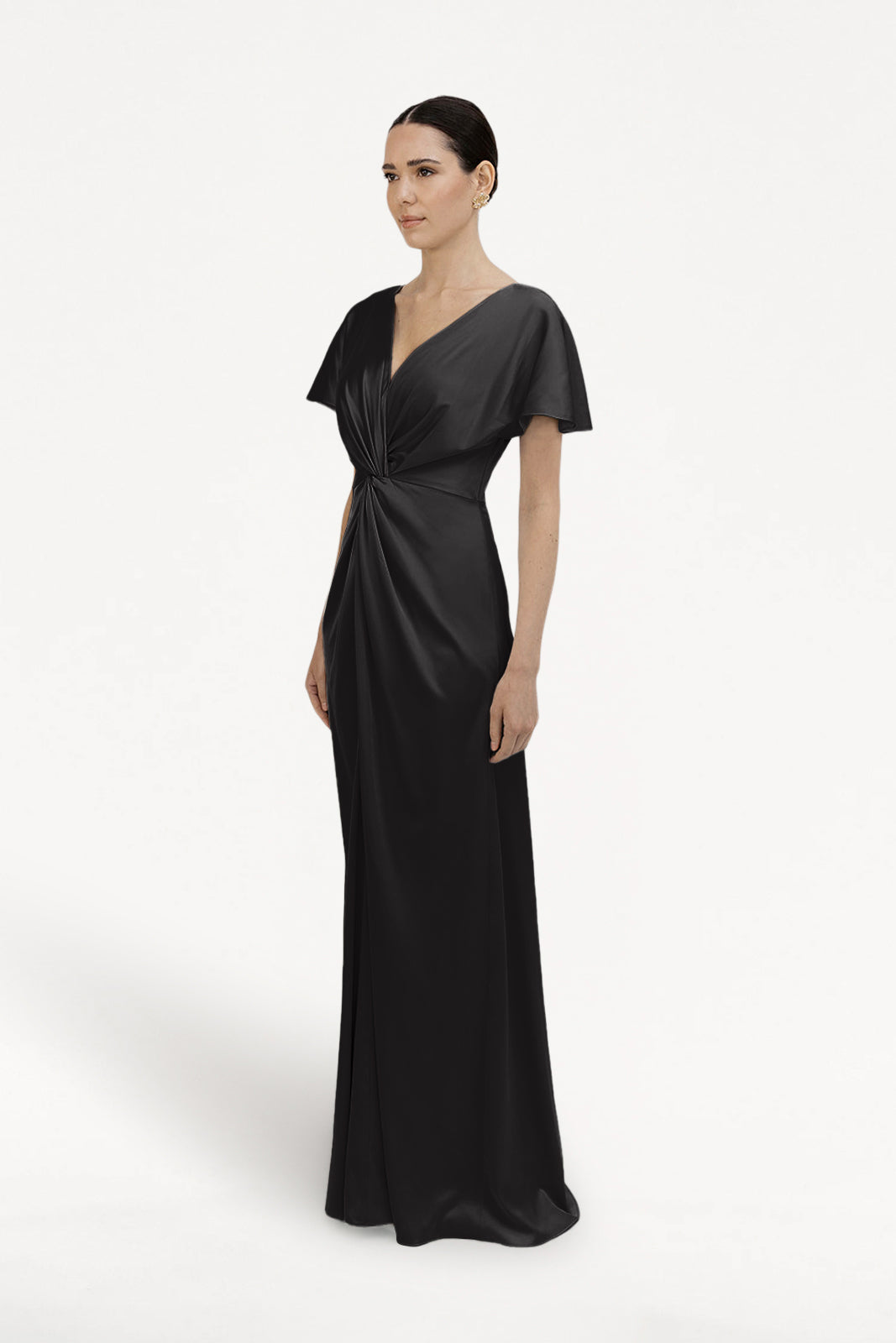 A woman with dark hair pulled back and a neutral expression wears the TH&TH Camilla Bridesmaid Dress in Black Satin—a size-inclusive, floor-length gown featuring a twist-knot bodice—against a plain white background.