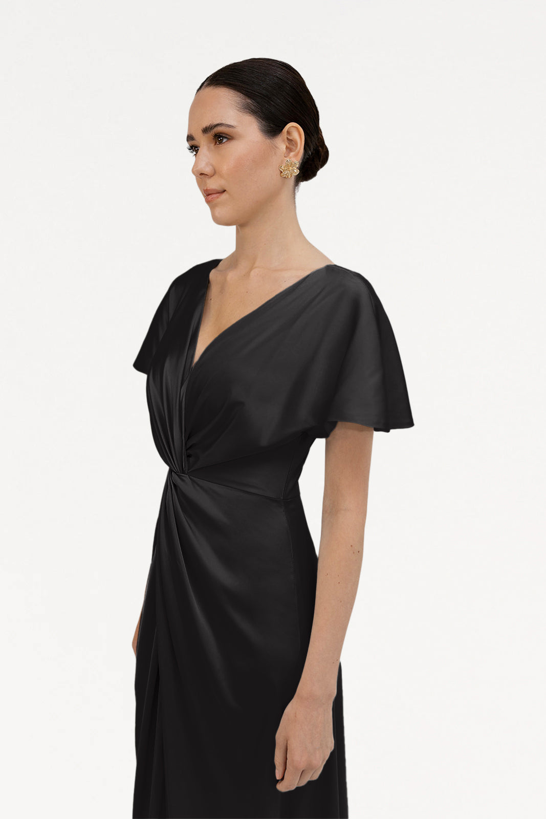 A woman models the size-inclusive TH&TH Camilla Bridesmaid Dress in Black Satin with flutter sleeves, V-neckline, and twist-knot bodice. She wears gold floral earrings and a sleek bun, photographed against a white background.