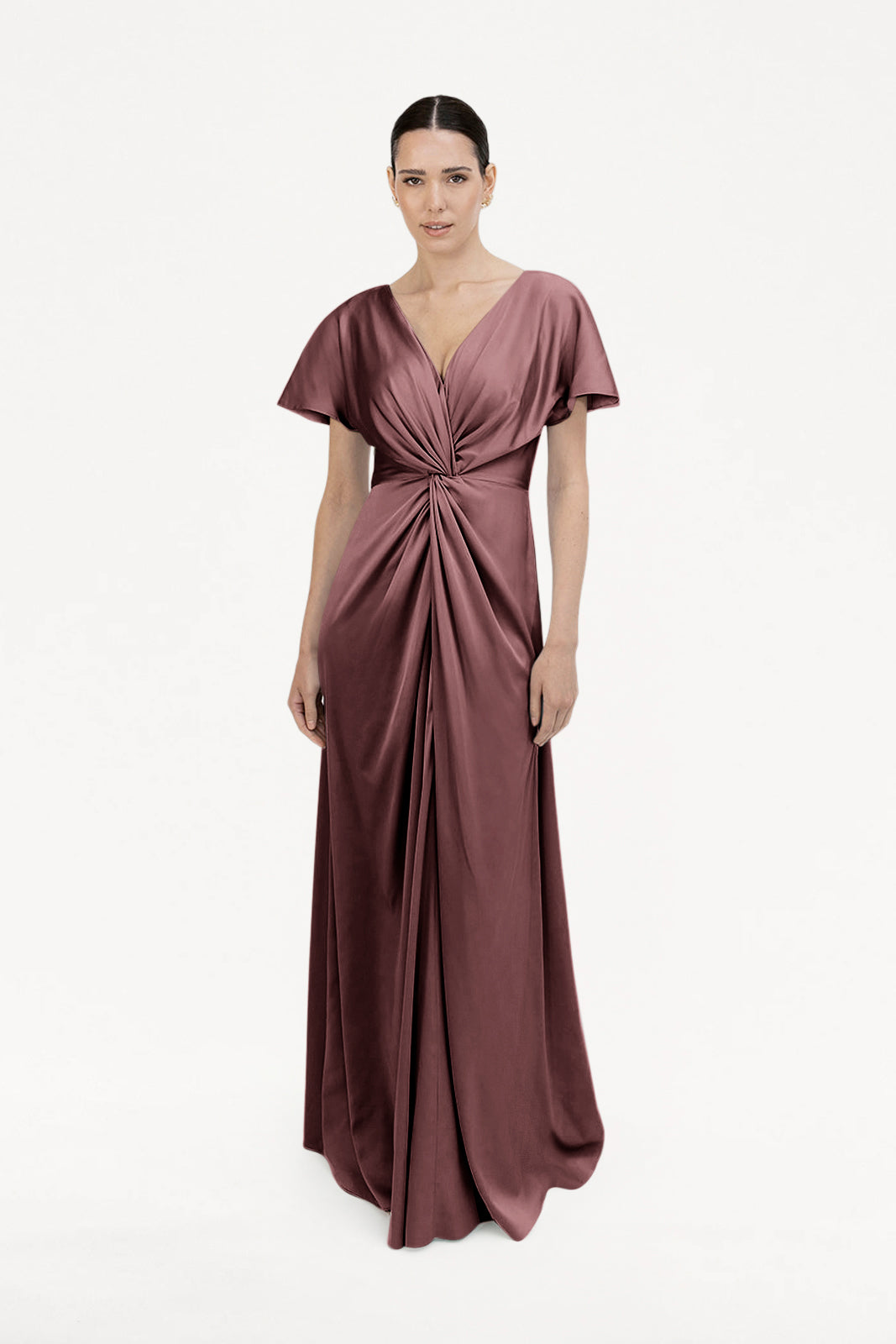 A woman stands against a white background wearing the Camilla Bridesmaid Dress in chocolate brown satin by TH&TH—a floor-length gown with short sleeves, a deep V-neckline, and a gathered twist waist, perfect for modern celebrations.