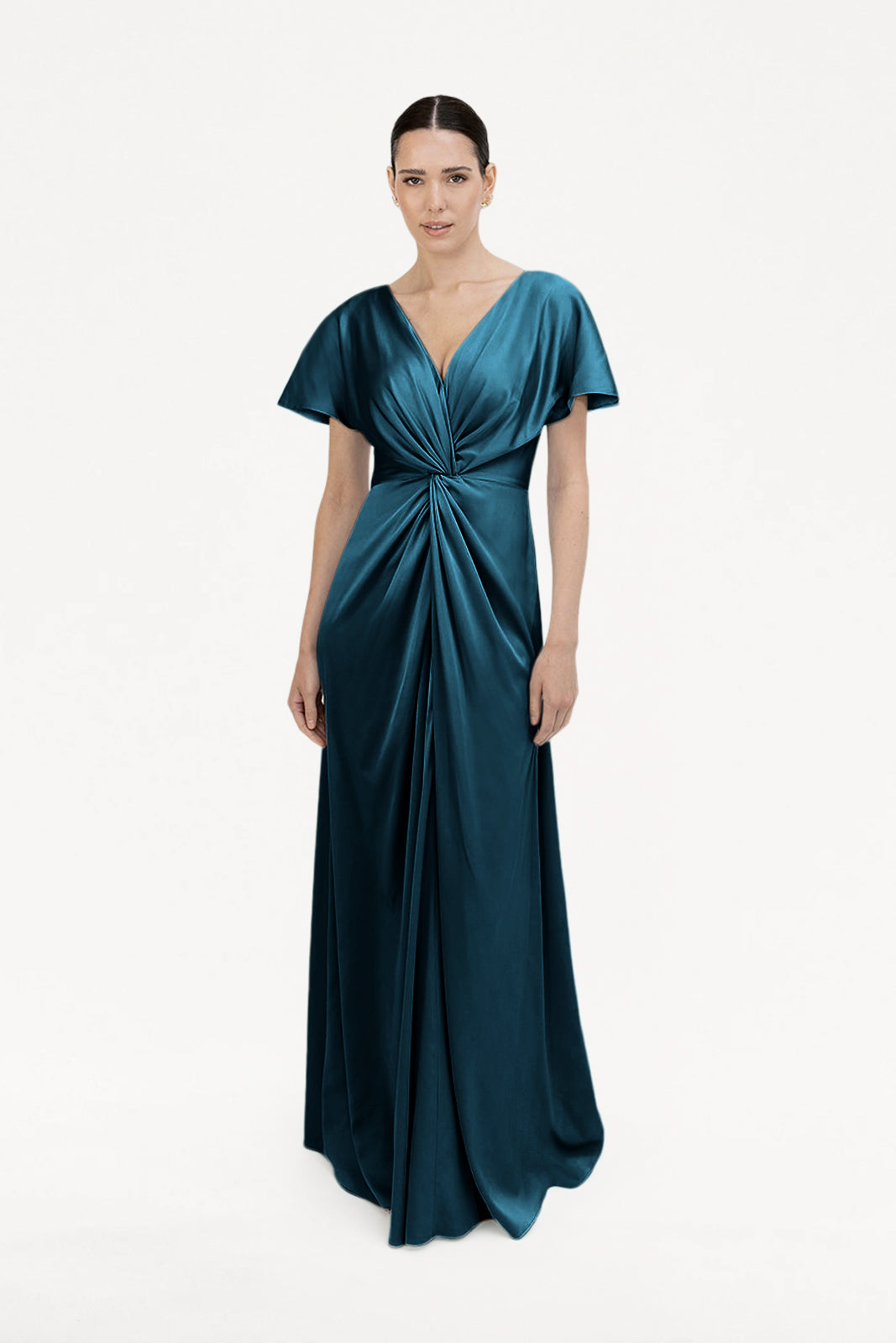 A woman models the Camilla Bridesmaid Dress in Emerald Dawn Satin by TH&TH, a floor-length gown featuring short sleeves, a deep V-neckline, and a gathered twist waist detail, set against a plain white background.