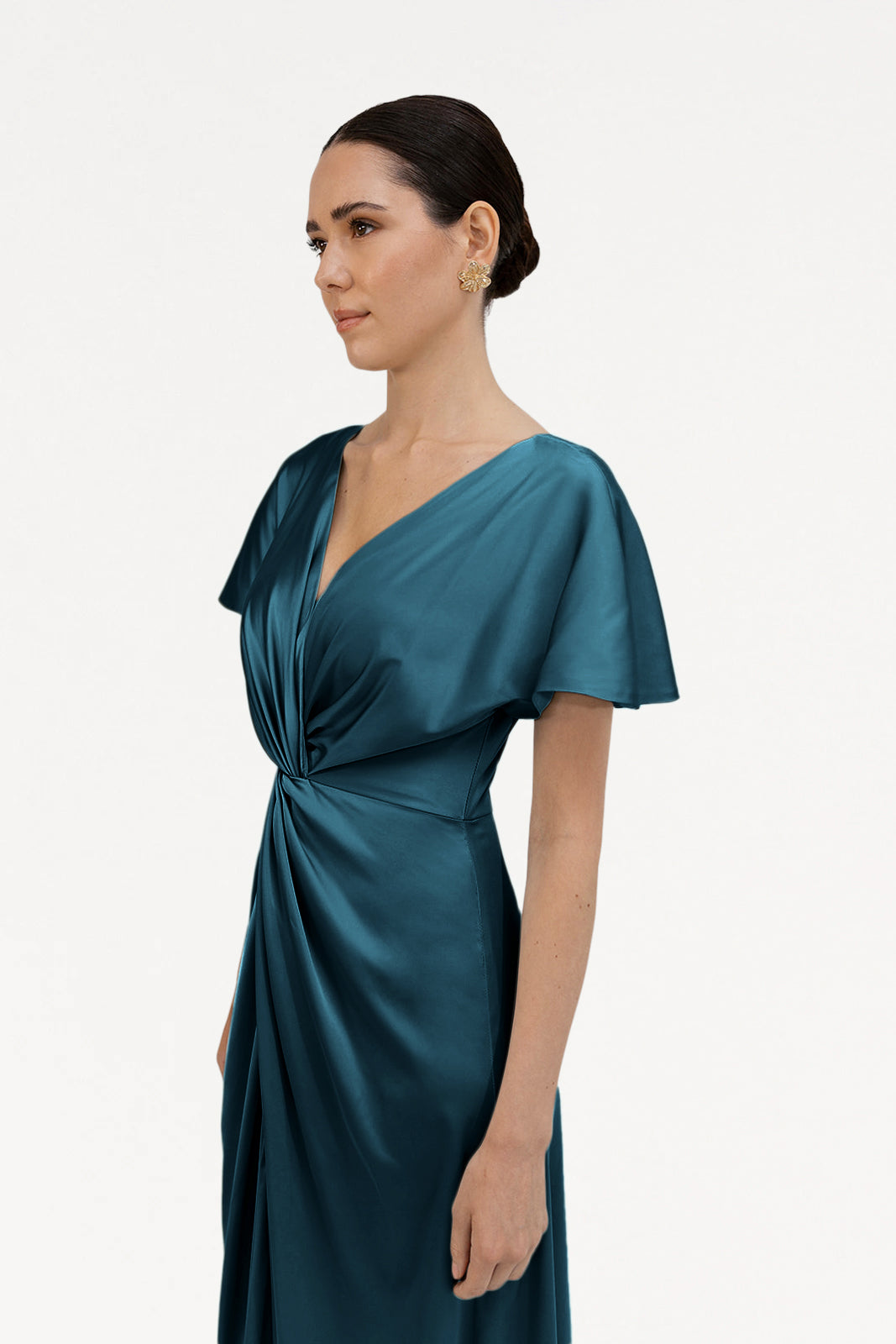 A woman with dark hair styled back models the TH&TH Camilla Bridesmaid Dress in Emerald Dawn Satin, a size-inclusive design with short sleeves, V-neck, and twisted front detail. She stands in profile against a plain white background.