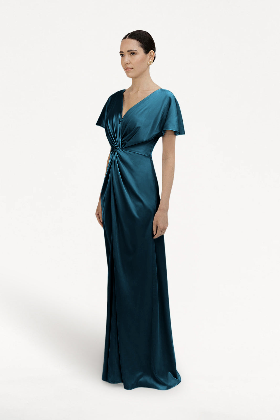 A woman poses against a white background in the Camilla Bridesmaid Dress by TH&TH, featuring emerald dawn satin, short sleeves, and a gathered twist waist detail. Her hair is styled in a sleek low bun for a modern bridesmaid look.