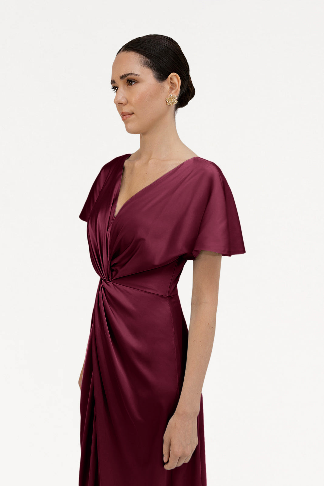 A woman with dark hair in a low bun wears the TH&TH Camilla Bridesmaid Dress in Merlot Satin, featuring short sleeves and a waist knot, standing against a plain white background.