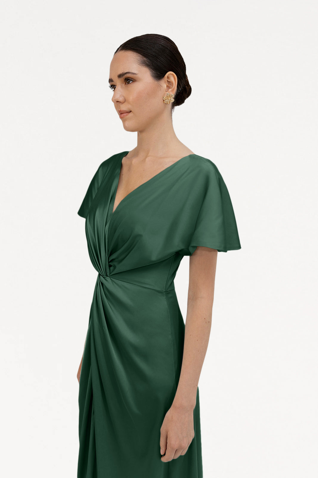 A woman with dark hair styled back wears the Camilla Bridesmaid Dress in Olive Grove Satin by TH&TH, featuring short sleeves, a V-neck, and a twisted knot waist detail, standing against a plain white background.