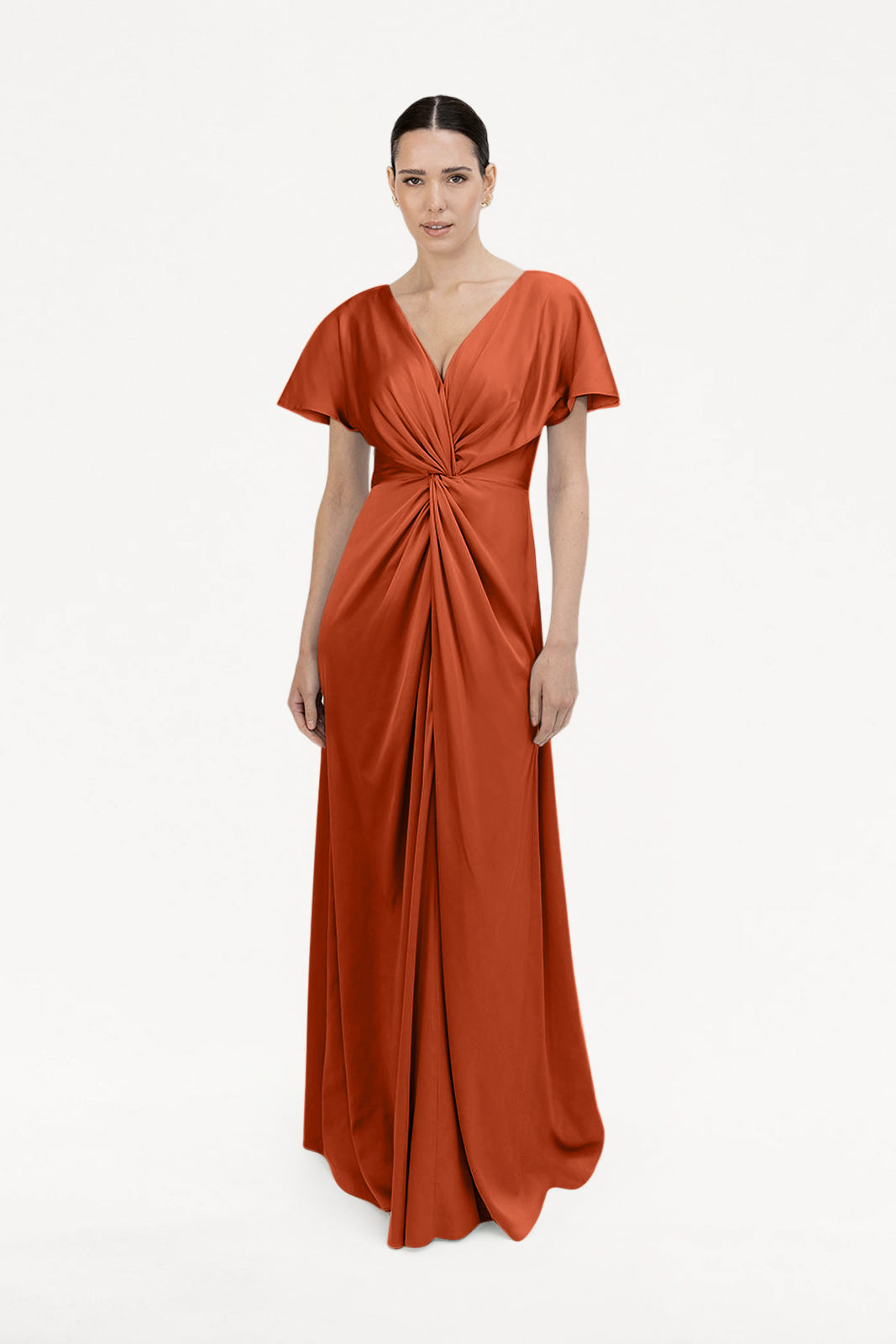 A woman wears the TH&TH Camilla Bridesmaid Dress in Terracotta Satin—a floor-length, short-sleeved, size-inclusive gown with a deep V-neck and twisted waist detail—posing against a plain white background.