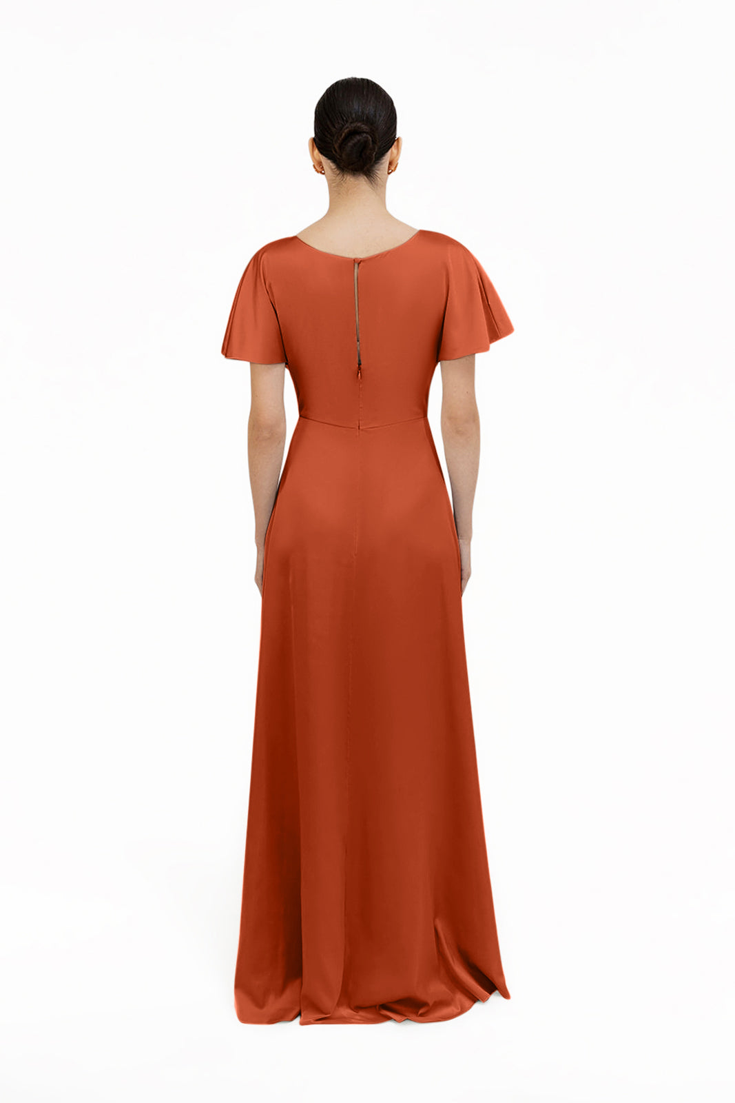 A woman with dark hair in a low bun models the floor-length, short-sleeved Camilla Bridesmaid Dress in Terracotta Satin by TH&TH, featuring a twist-knot bodice, shown from the back against a plain white background.
