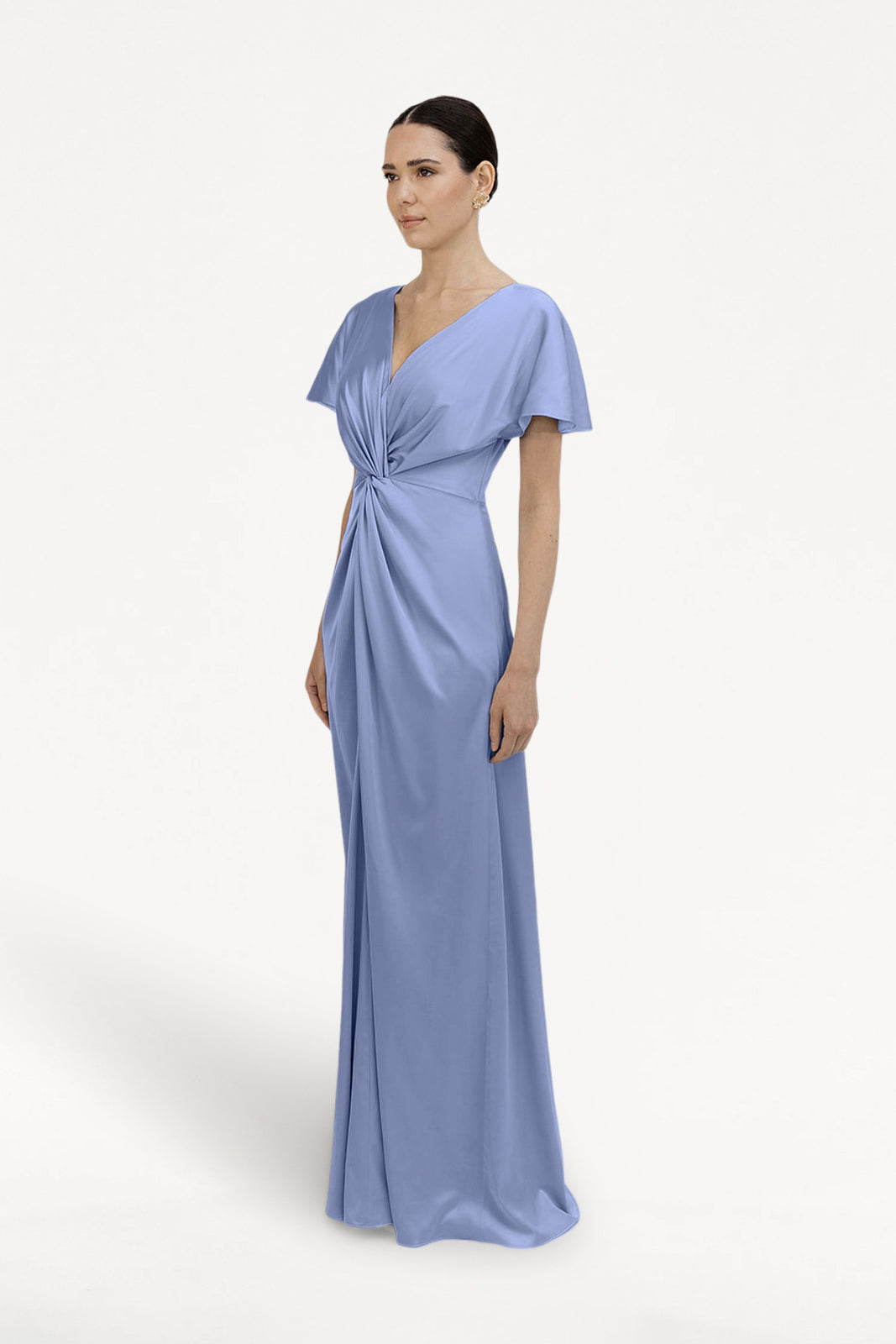 A woman with dark hair pulled back models the TH&TH Camilla Bridesmaid Dress in French Blue Satin, featuring a floor-length skirt, short sleeves, V-neckline, and twist-knot bodice, set against a plain white background.