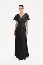 A woman poses against a white background in the TH&TH Camilla Bridesmaid Dress in Black Satin, a floor-length gown with short sleeves, a twist-knot bodice, and deep V-neckline. Her dark hair is styled in a sleek low bun.