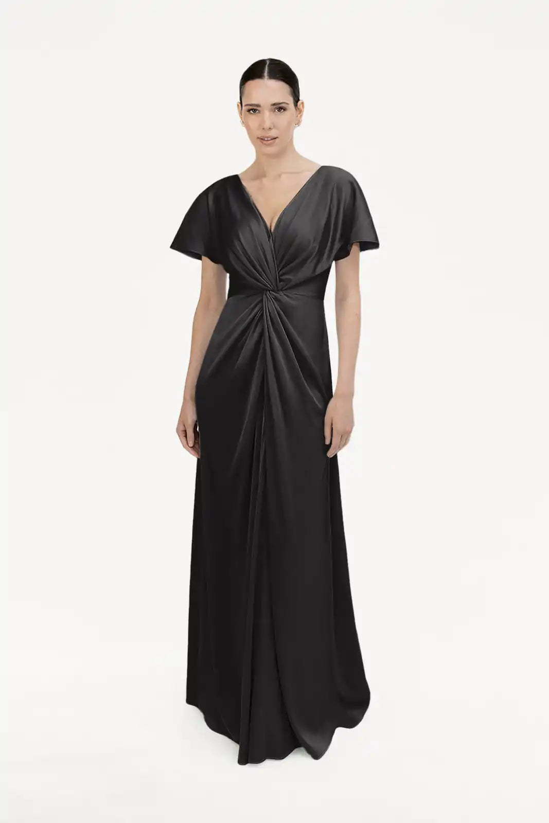 A woman poses against a white background in the TH&TH Camilla Bridesmaid Dress in Black Satin, a floor-length gown with short sleeves, a twist-knot bodice, and deep V-neckline. Her dark hair is styled in a sleek low bun.