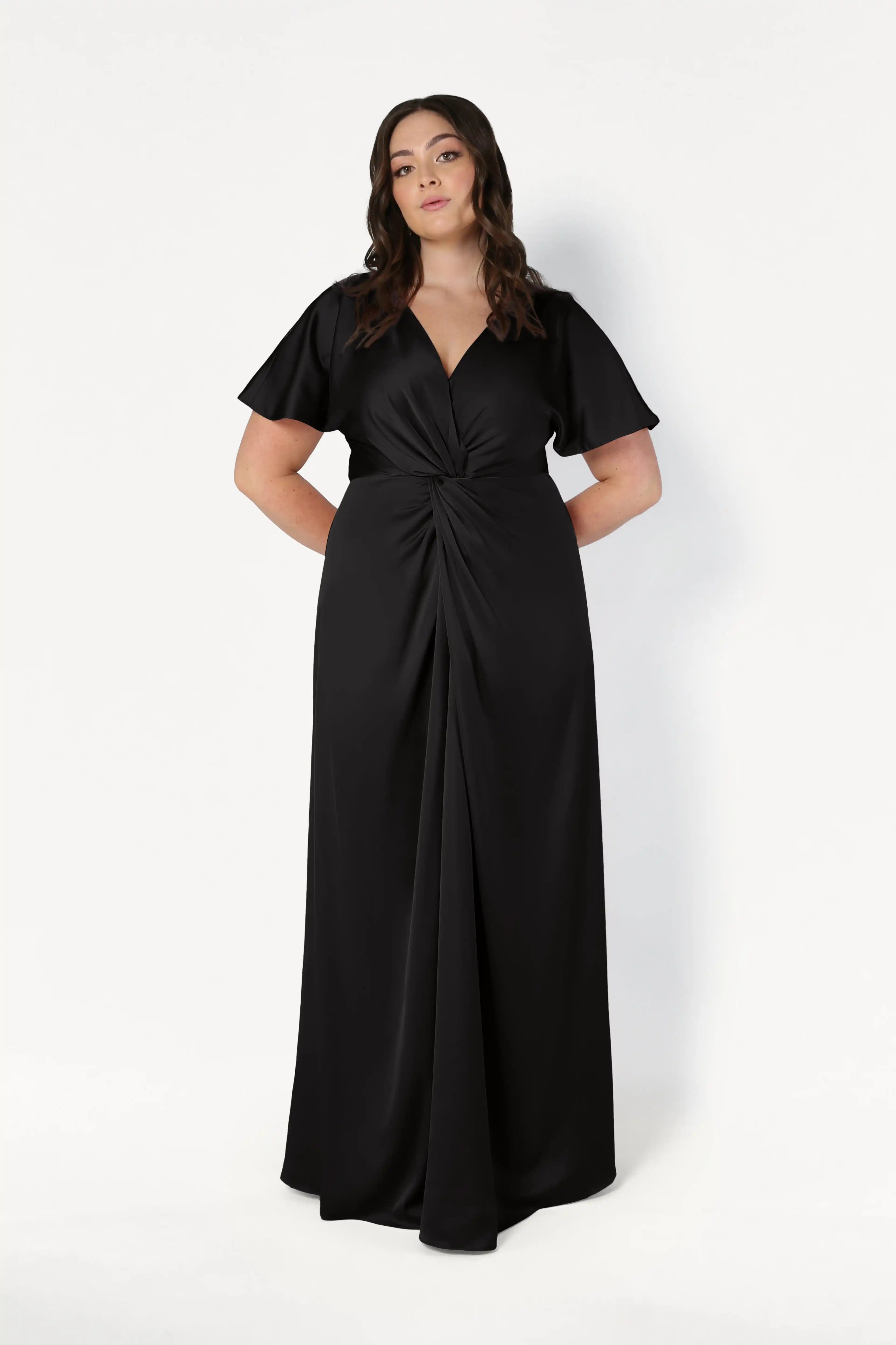 A woman models the Camilla Bridesmaid Dress in Black Satin by TH&TH, featuring a floor-length silhouette, short sleeves, V-neckline, and twist-knot bodice, standing confidently against a white background.