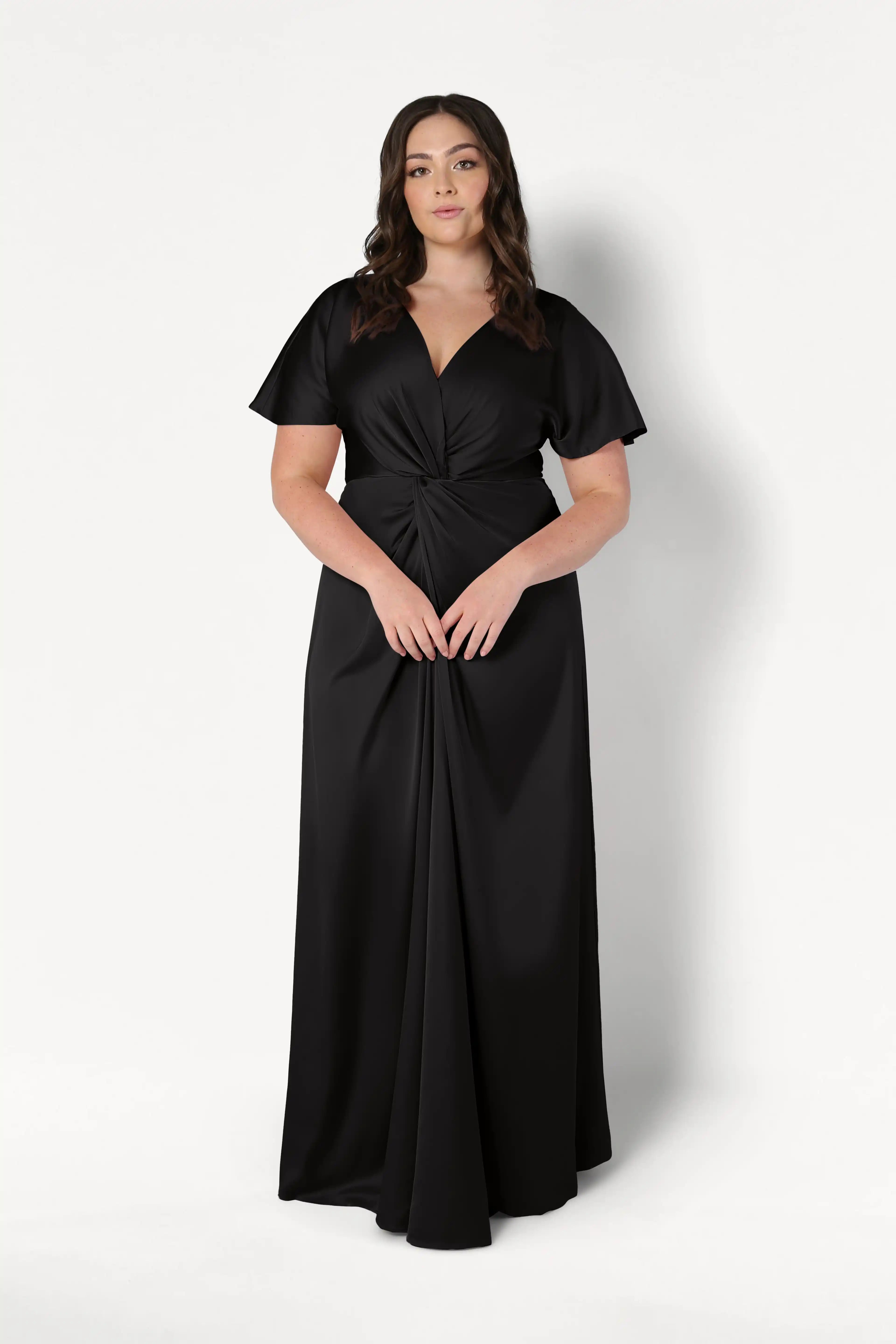 A woman wears the Camilla Bridesmaid Dress in Black Satin by TH&TH—a floor-length, short-sleeved satin gown with a twist-knot bodice—standing against a plain white background.