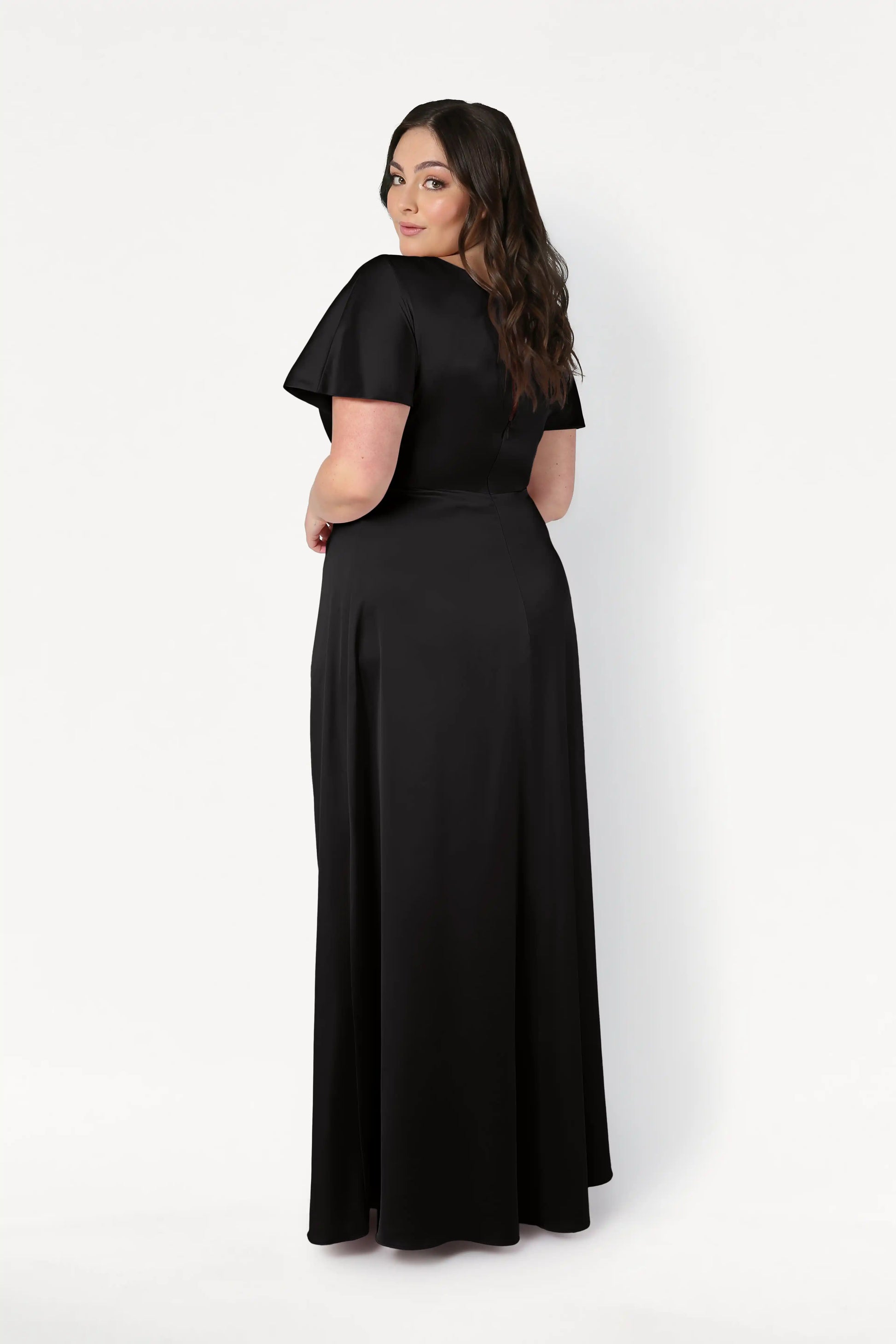A woman with long dark hair, facing away and looking over her shoulder, models the size-inclusive TH&TH Camilla Bridesmaid Dress in Black Satin—a floor-length gown with a twist-knot bodice—against a plain white background.