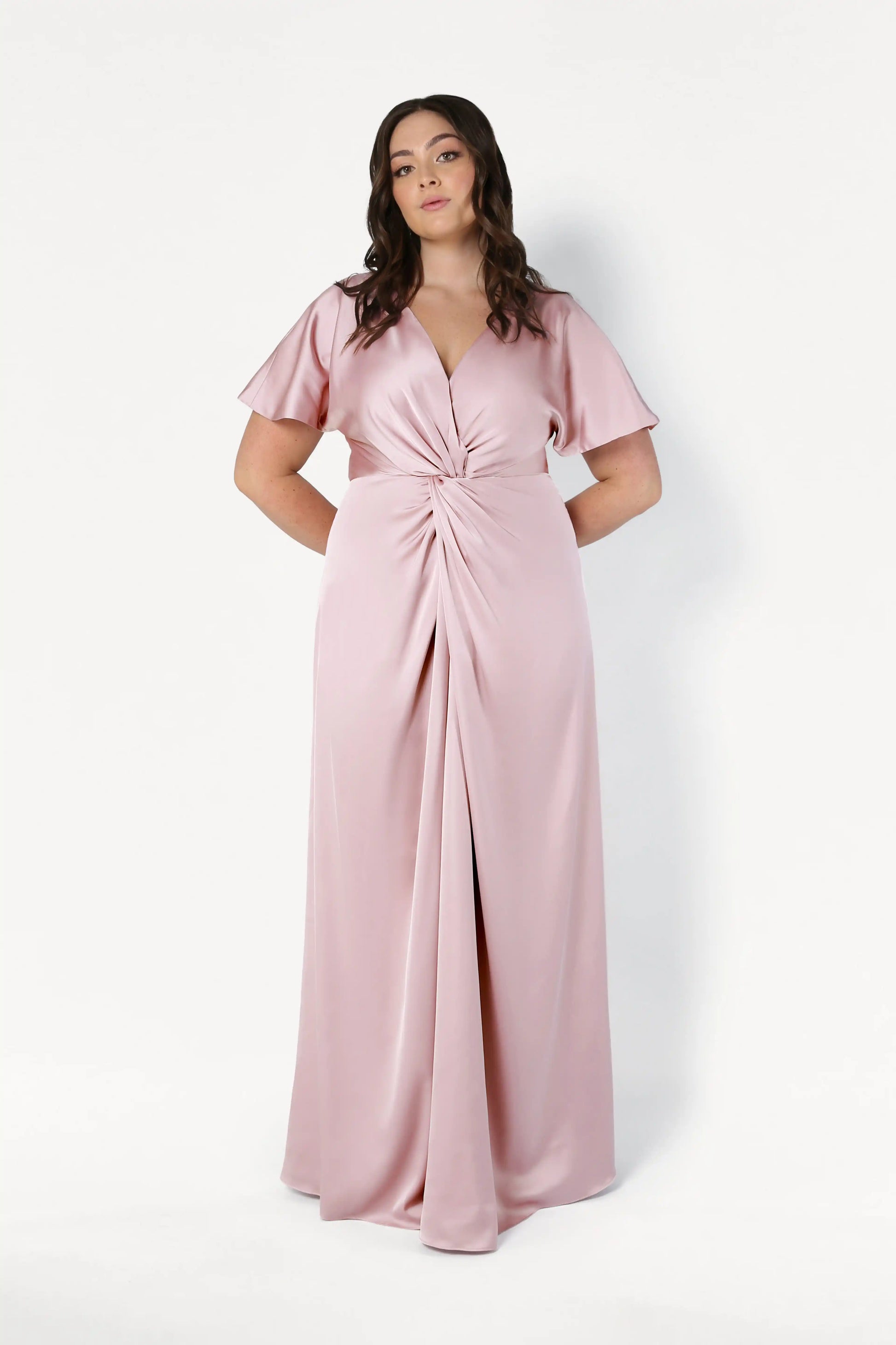 A woman stands against a plain white background wearing the TH&TH Camilla Bridesmaid Dress in blush pink satin, featuring short sleeves, a V-neckline, floor-length design, and a gathered twist detail at the waist.