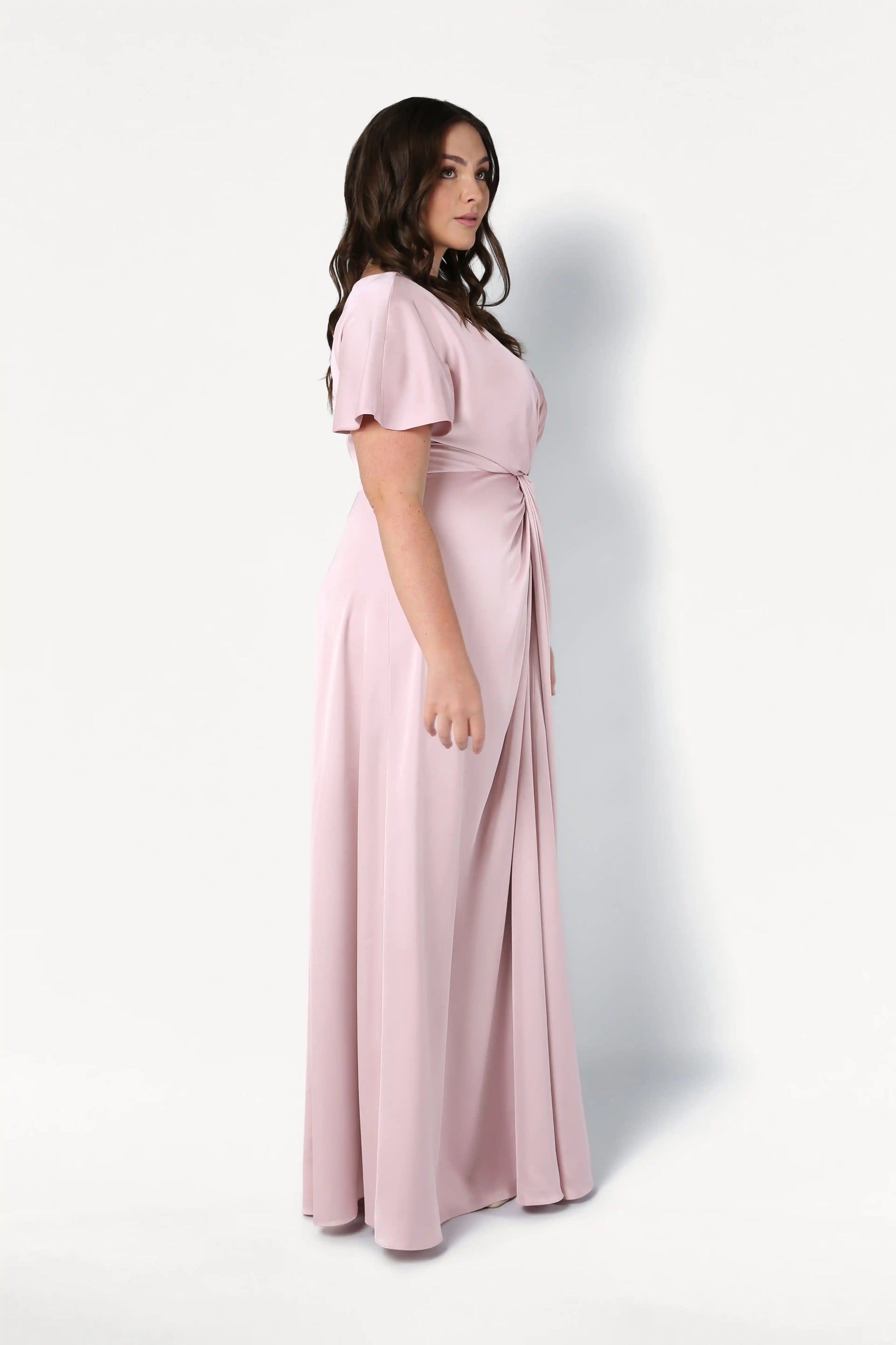 A woman with long brown hair poses sideways in the TH&TH Camilla Bridesmaid Dress in blush pink satin—a size-inclusive, floor-length gown with short sleeves and gathered waist—against a plain white background.