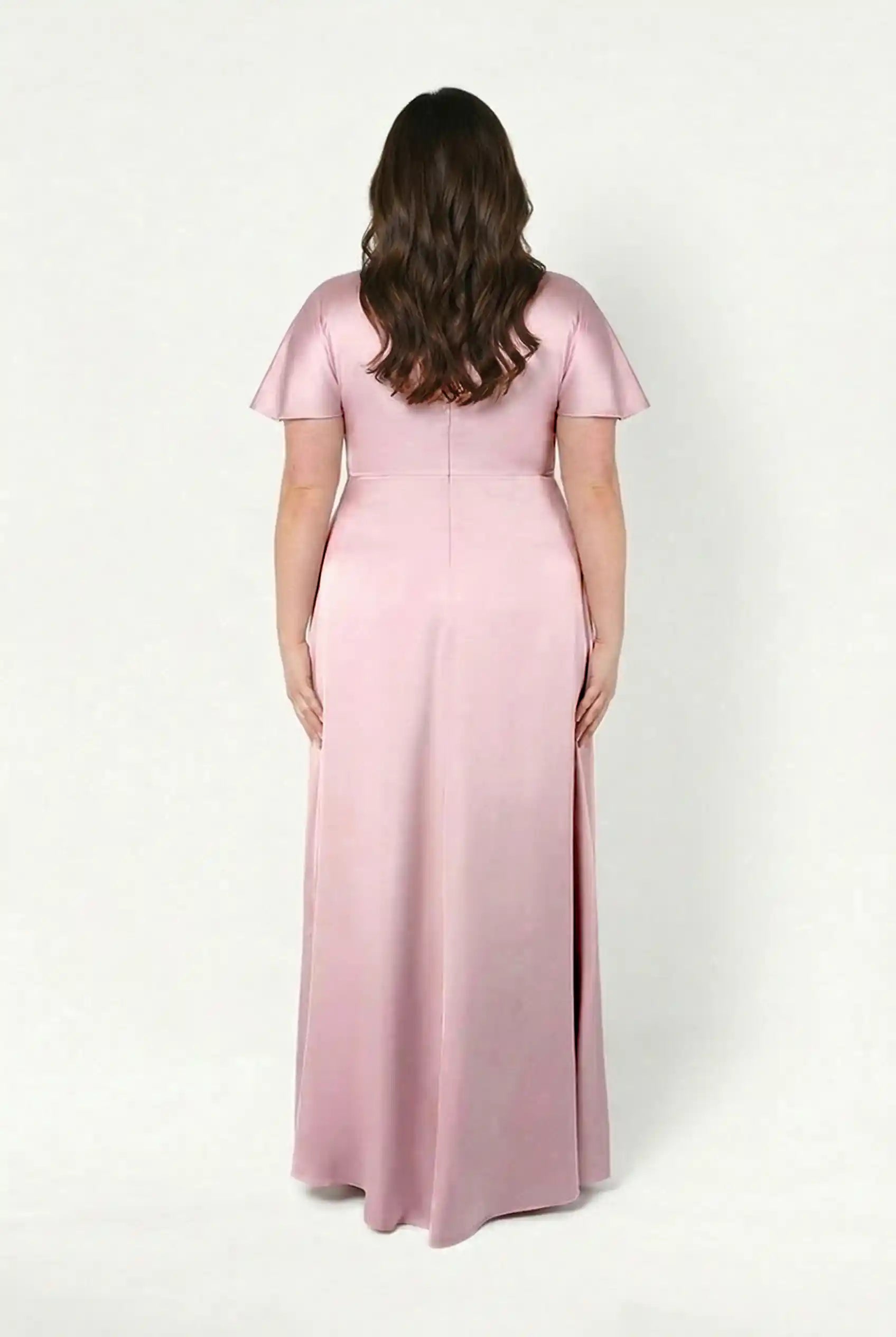 A woman with long brown hair, seen from the back, wears the Camilla Bridesmaid Dress in blush pink satin by TH&TH—a floor-length, short-sleeved modern gown—set against a plain white background.