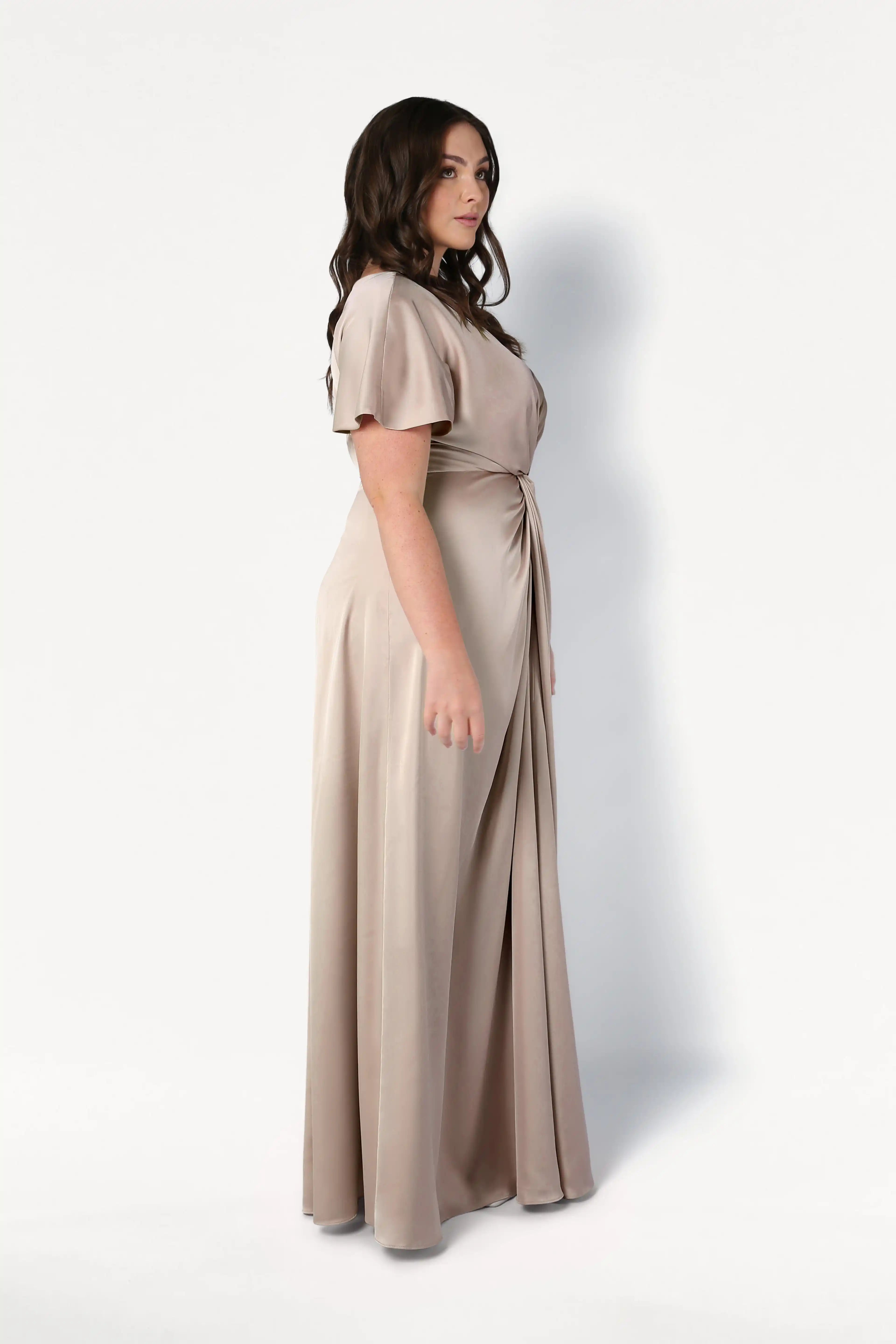 A woman with long dark hair stands in profile, wearing the TH&TH Camilla Bridesmaid Dress in Champagne Sky Satin—a size-inclusive, floor-length gown with short sleeves and a gathered waist—against a plain white background.