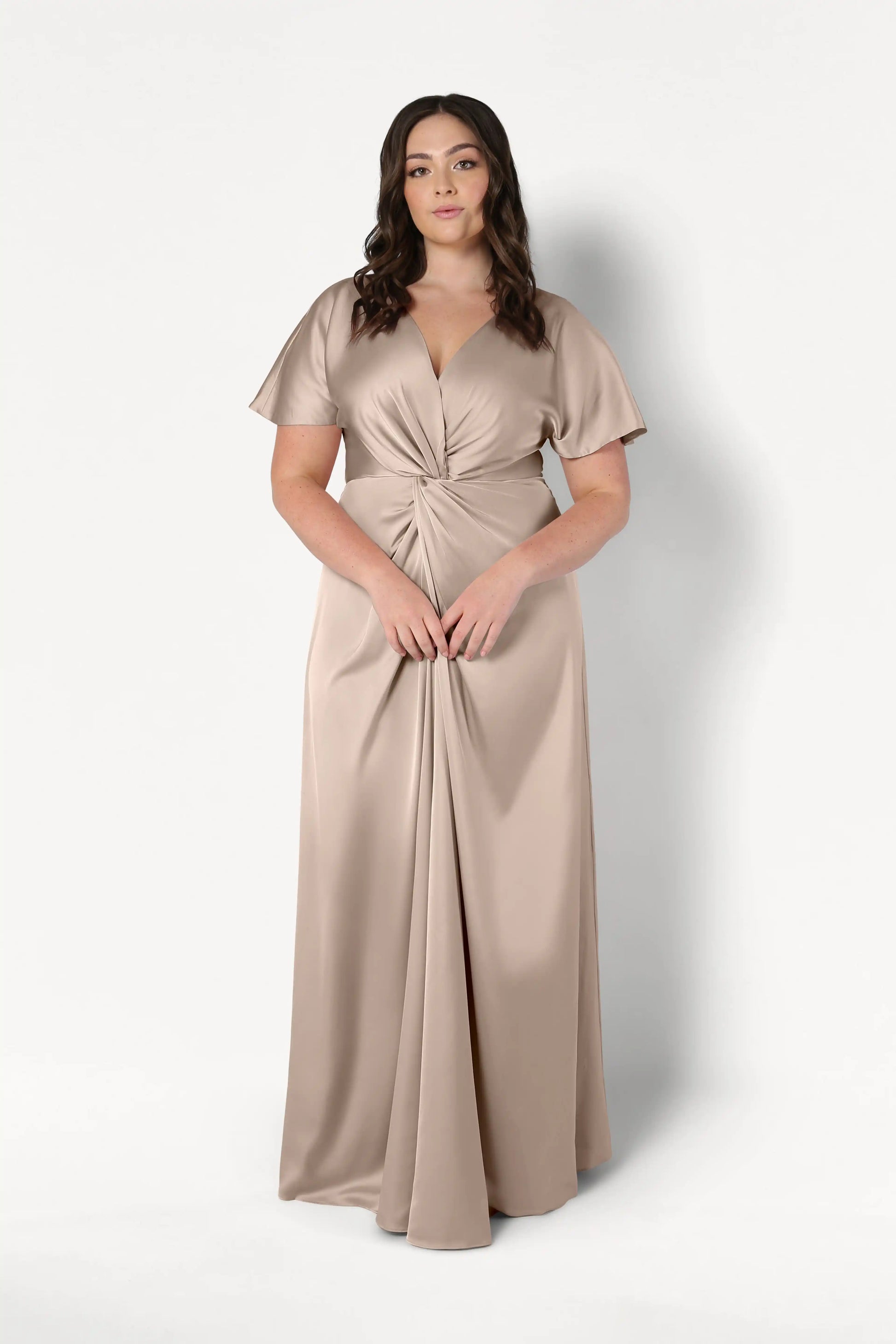 A woman with long wavy brown hair models the Camilla Bridesmaid Dress in Champagne Sky Satin by TH&TH, a floor-length gown with a twist-knot bodice and V-neckline, standing against a plain white background.