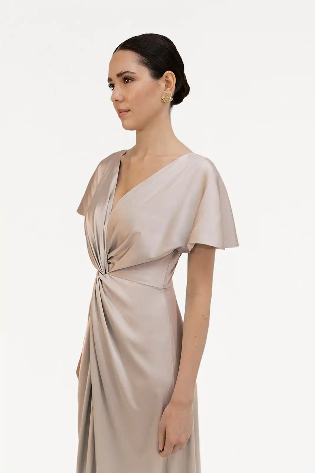A woman with dark hair in a low bun wears the TH&TH Camilla Bridesmaid Dress in Champagne Sky Satin, a size-inclusive gown with a twist-knot bodice and short sleeves, standing against a plain white background.