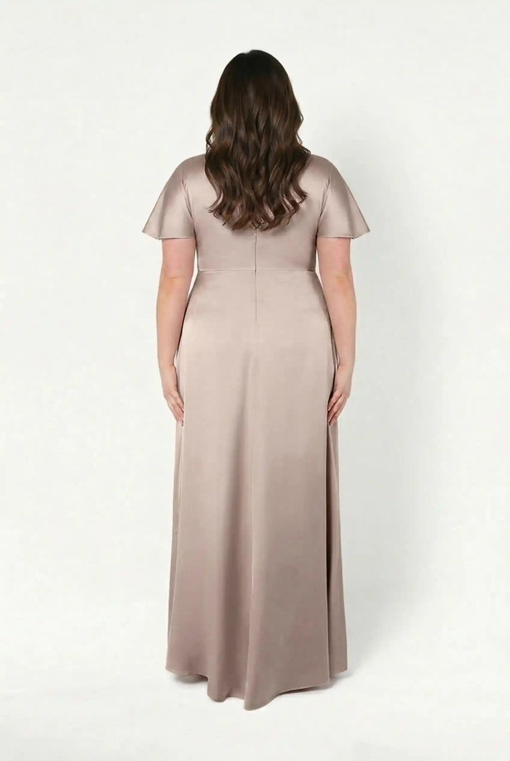 A woman with long brown hair faces away, wearing the Camilla Bridesmaid Dress in Champagne Sky Satin by TH&TH—a floor-length, short-sleeve gown with a twist-knot bodice—against a plain white background.