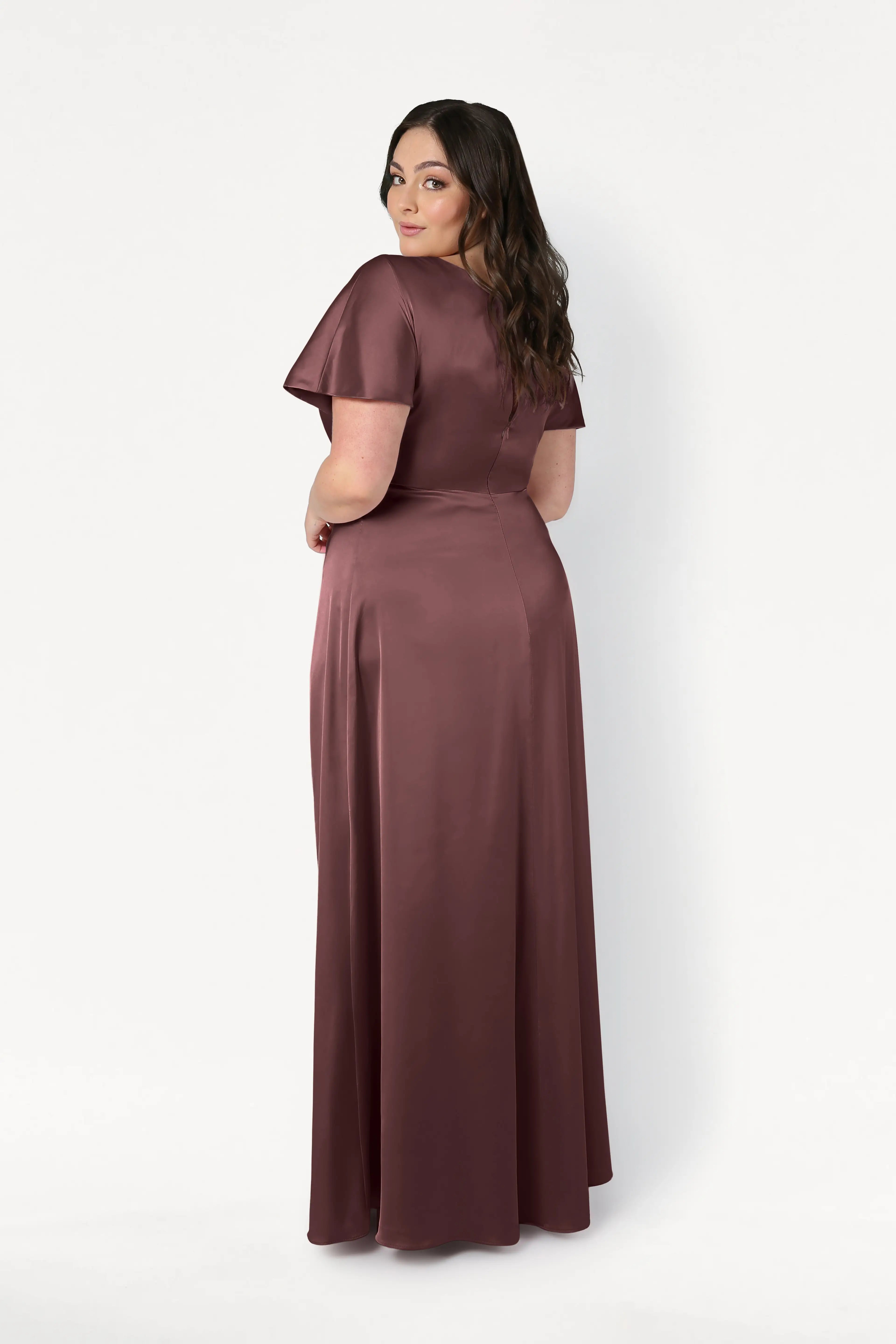 A woman with long brown hair stands facing backward, looking over her shoulder. She wears the TH&TH Camilla Bridesmaid Dress in chocolate brown satin against a plain white background.