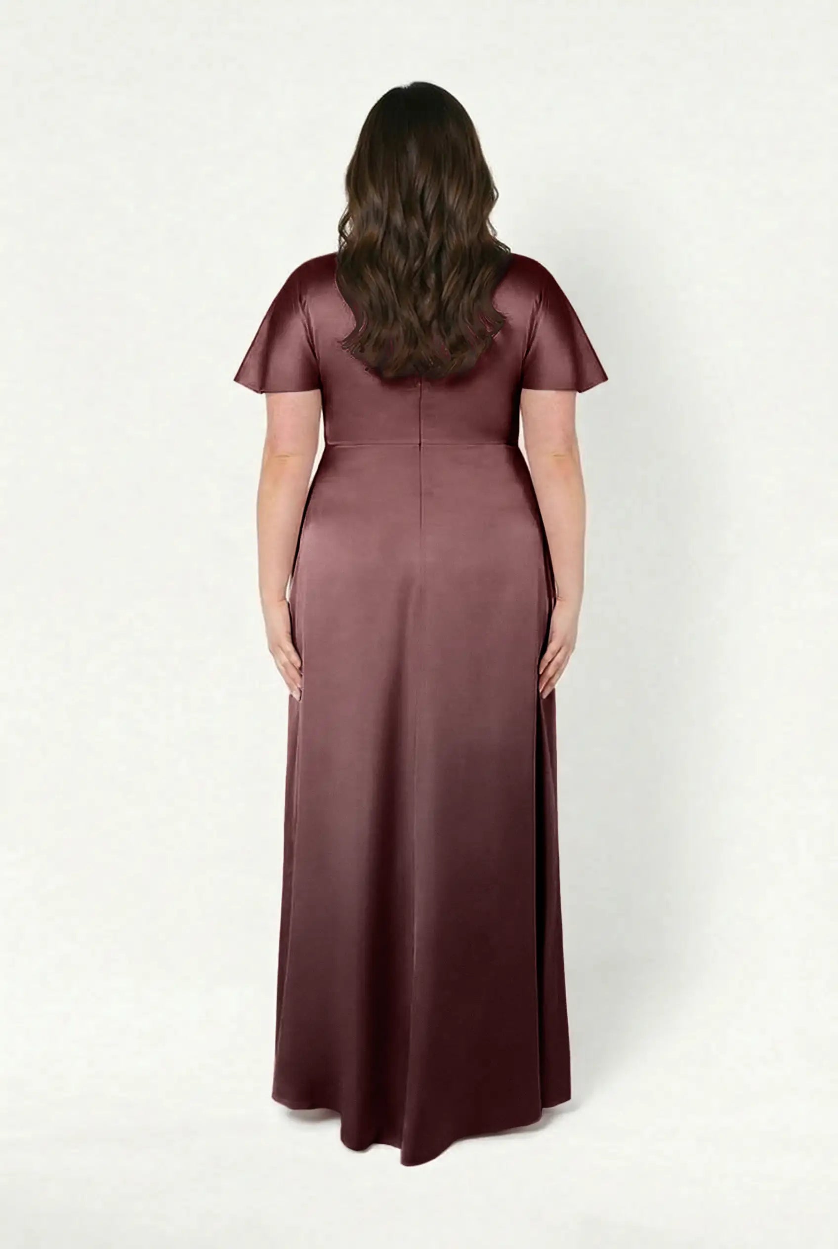 A woman with long brown hair stands facing away, wearing the Camilla Bridesmaid Dress in Chocolate Brown Satin by TH&TH—a floor-length, short-sleeved modern gown—against a plain light background.