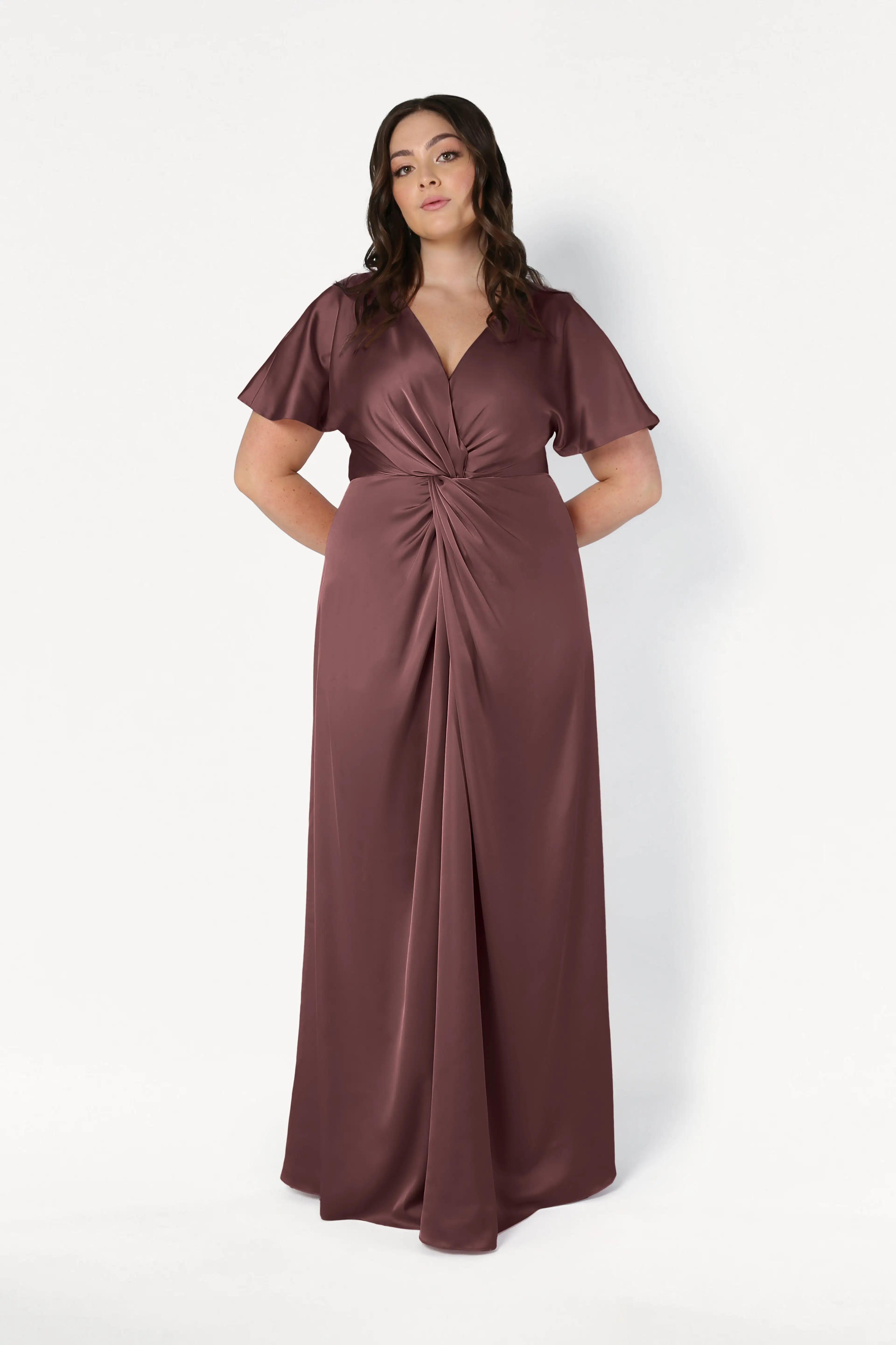 A woman wears the TH&TH Camilla Bridesmaid Dress in chocolate brown satin, featuring flutter sleeves, a V-neckline, and a gathered knot waist, standing against a white background. This modern dress exudes effortless style and elegance.