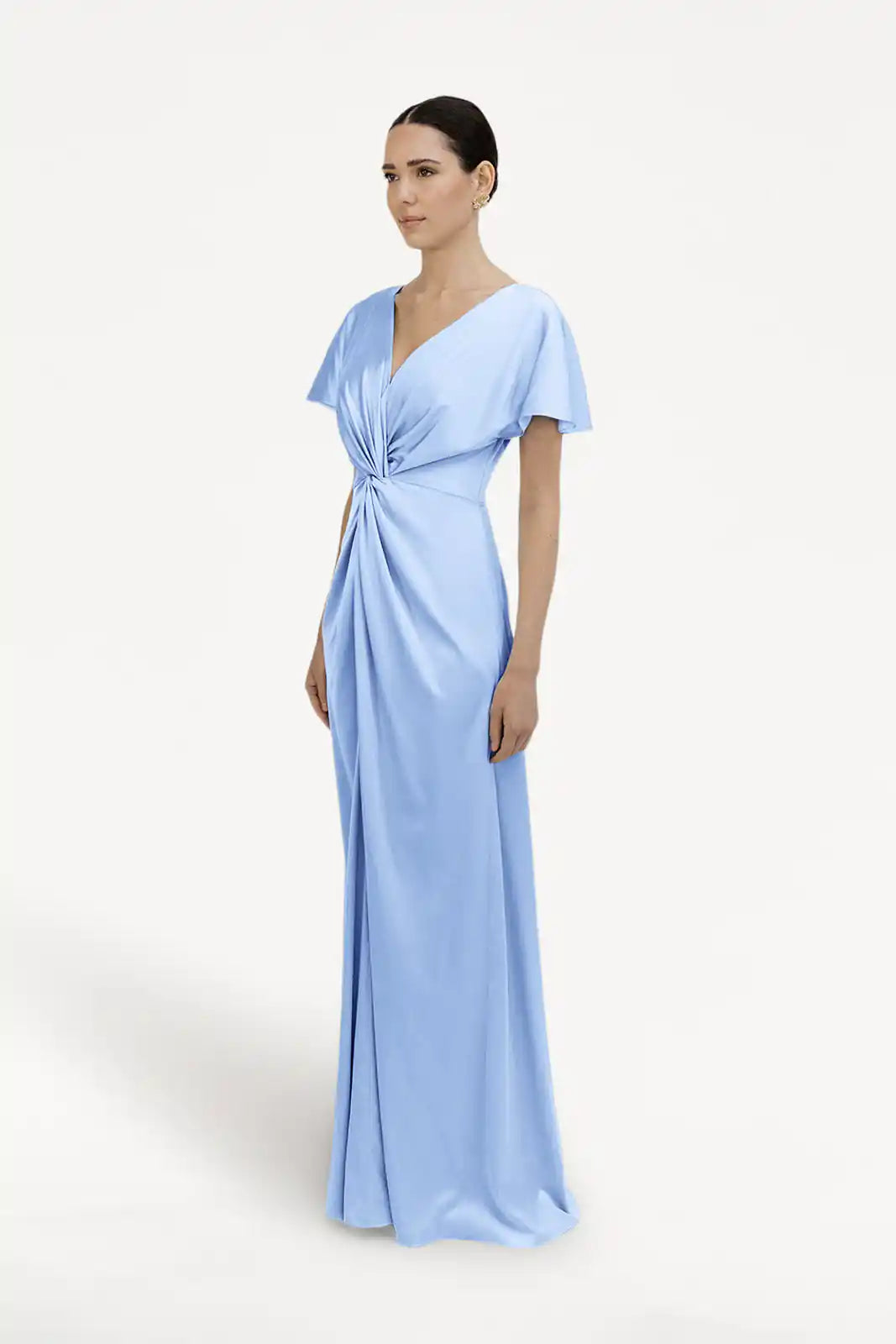A woman stands against a white background wearing the TH&TH Camilla Bridesmaid Dress in Dusty Blue Satin, a modern, floor-length gown with short sleeves, V-neckline, and a gathered twisted waist. Her hair is neatly pulled back.
