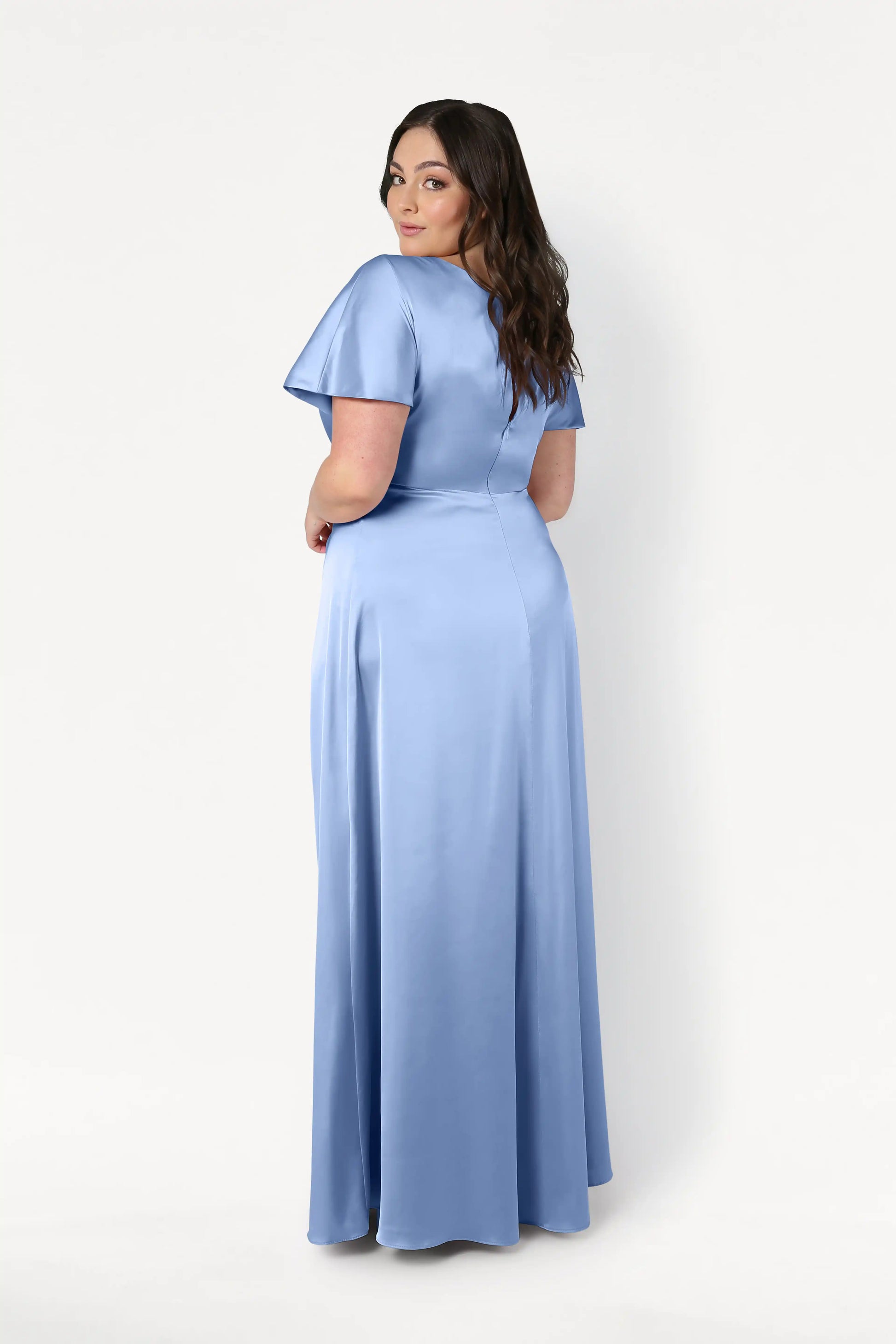 A woman with long brown hair models the Camilla Bridesmaid Dress in Dusty Blue Satin by TH&TH, featuring short sleeves and a floor-length design, as she looks over her shoulder against a plain white background.