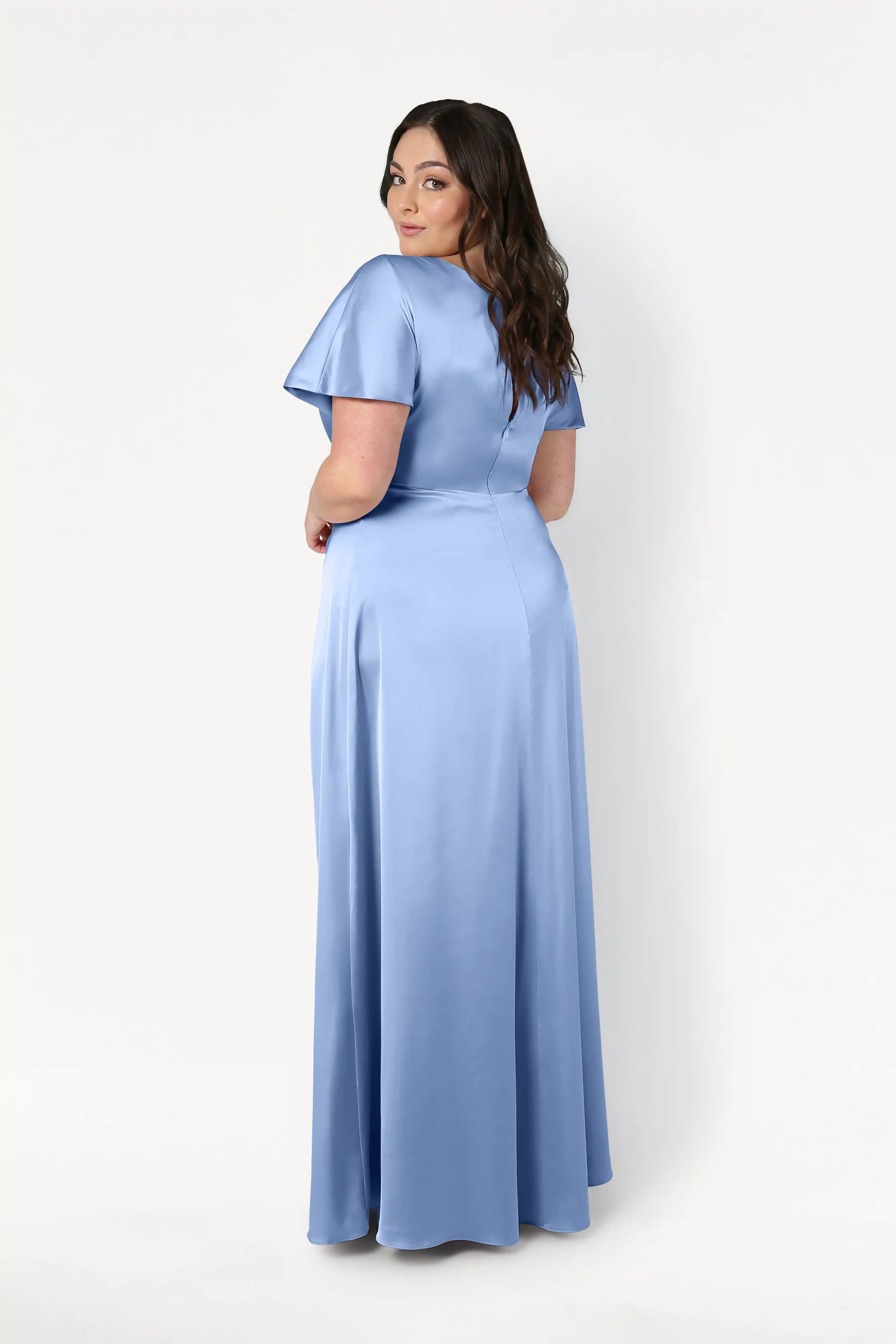 A woman with long brown hair models the Camilla Bridesmaid Dress in Dusty Blue Satin by TH&TH, featuring short sleeves and a floor-length design, as she looks over her shoulder against a plain white background.