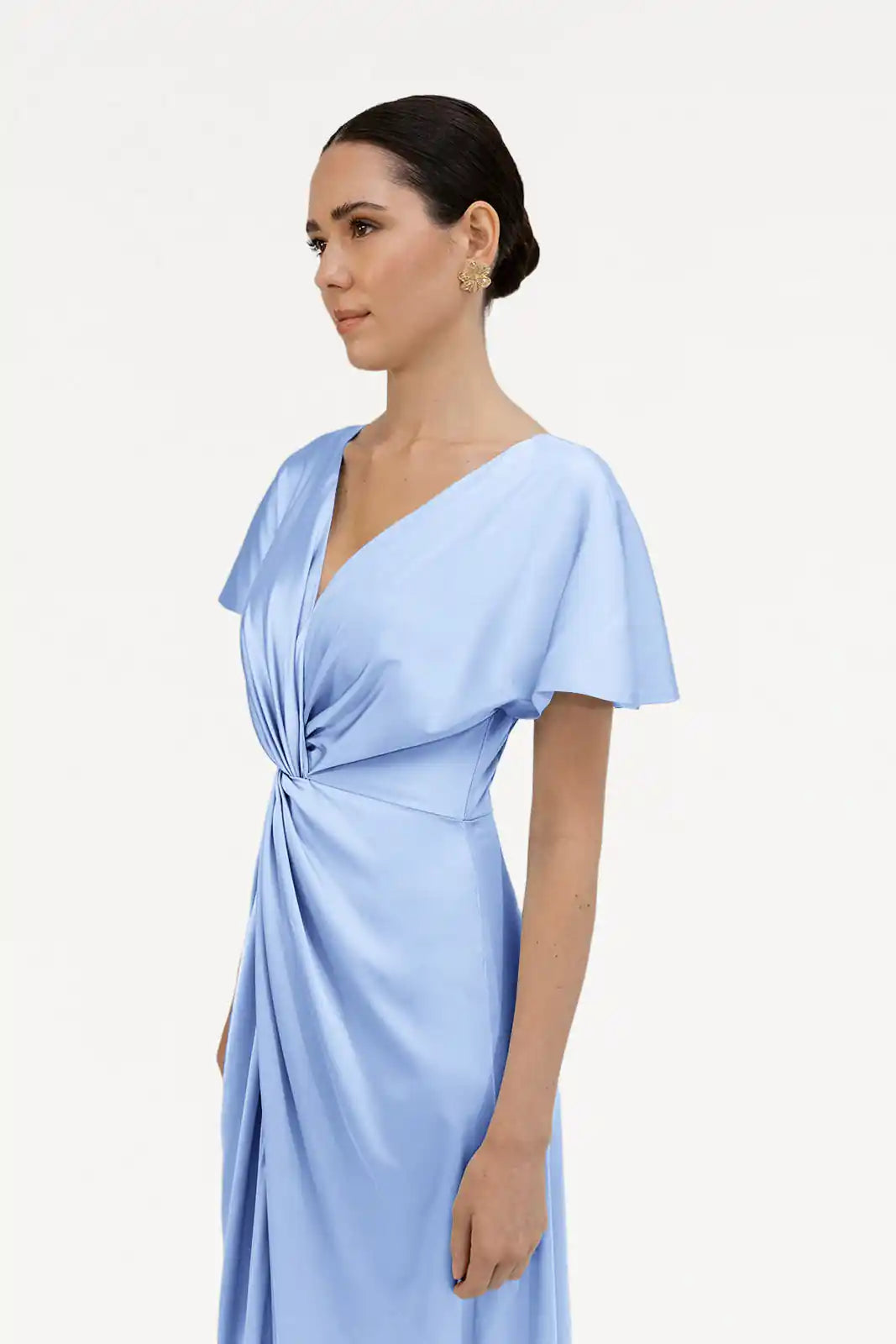 A woman with dark hair in a low bun wears the TH&TH Camilla Bridesmaid Dress in Dusty Blue Satin, featuring short sleeves, a V-neck, and a draped twist waist detail, standing before a plain white background.