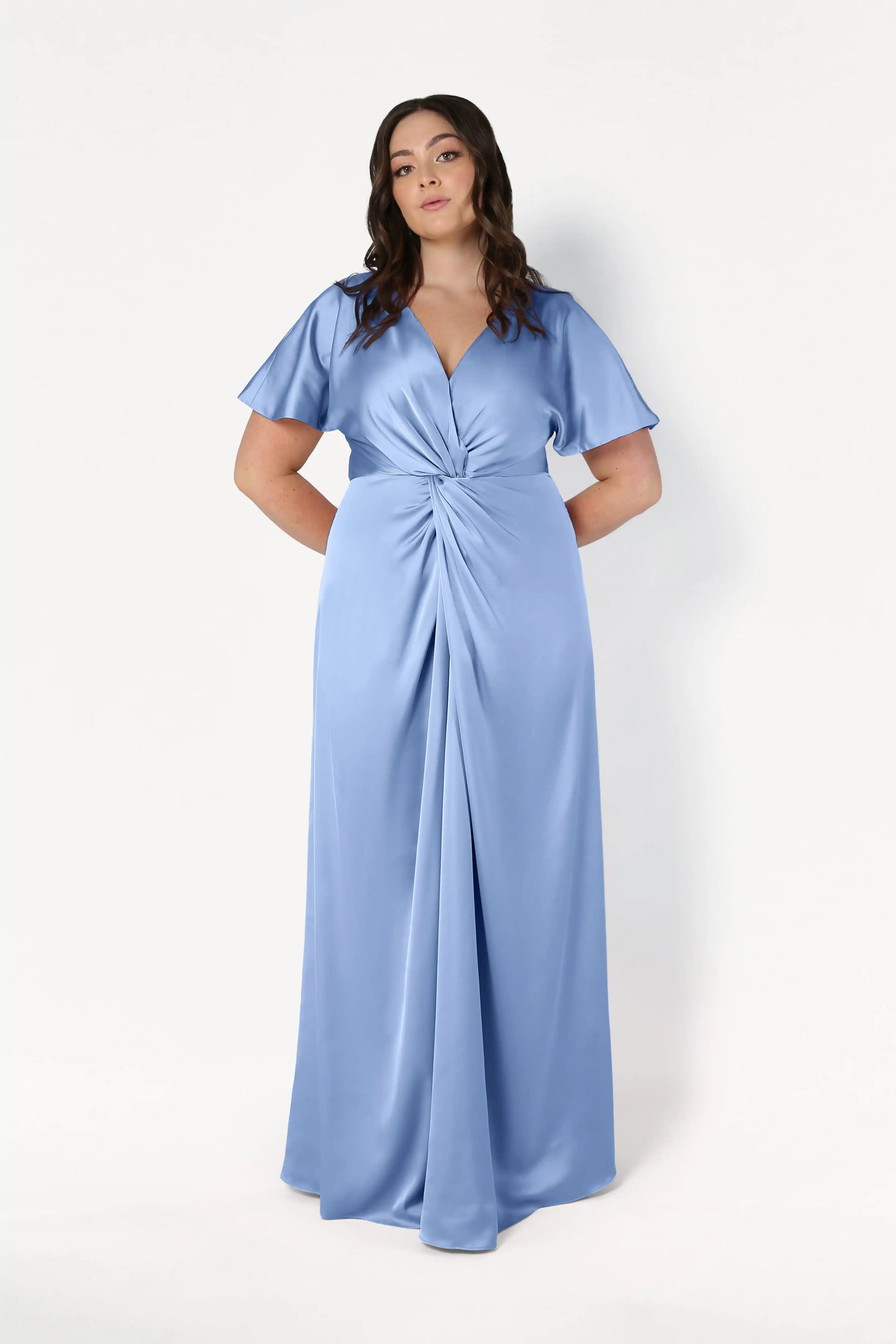 A woman models the TH&TH Camilla Bridesmaid Dress in Dusty Blue Satin, featuring flutter sleeves, a deep V-neckline, and a gathered knot waist, standing against a plain white background.