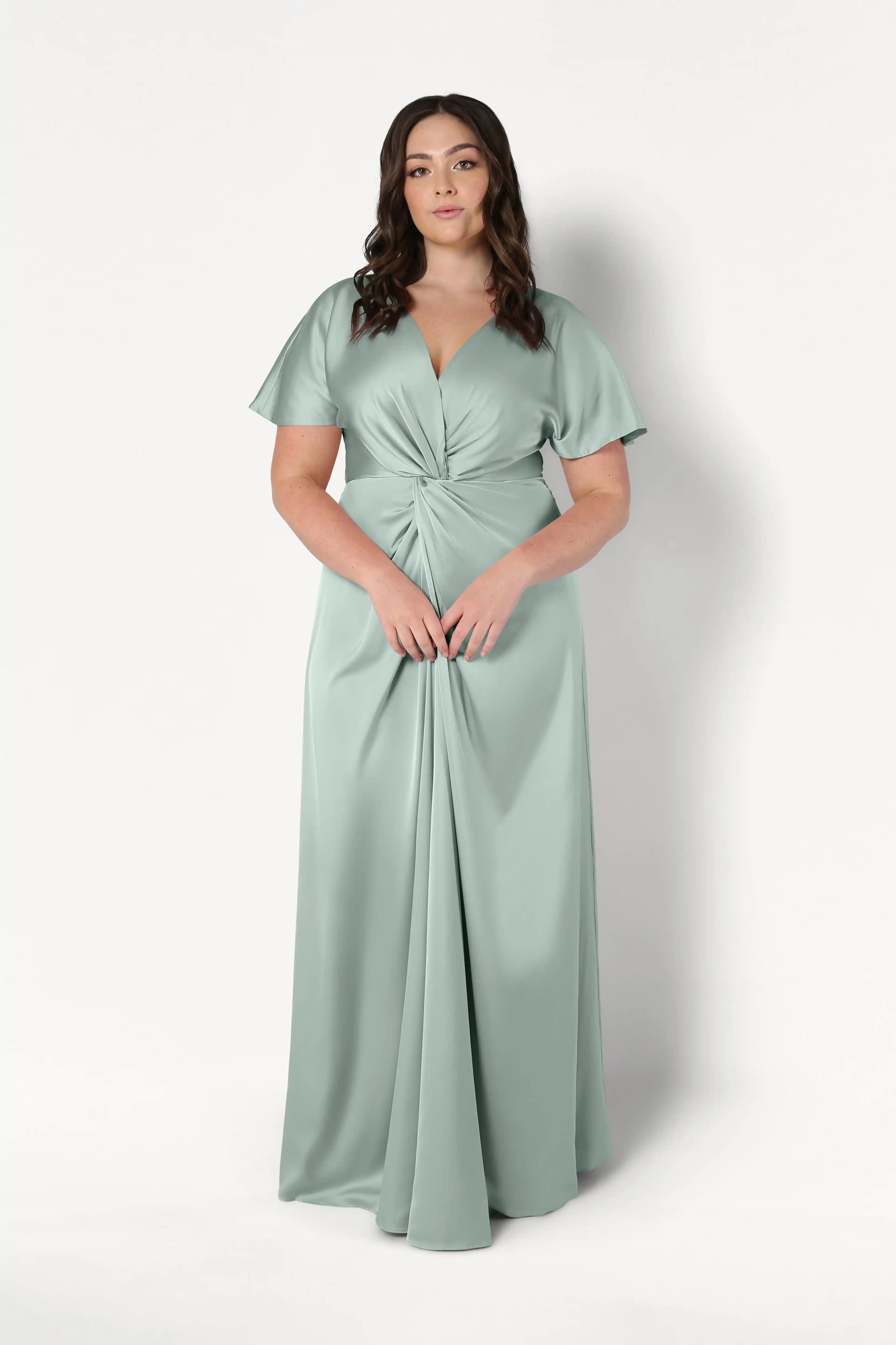 A woman poses against a white background wearing the TH&TH Camilla Bridesmaid Dress in Dusty Eucalyptus Satin, a floor-length gown with short flutter sleeves, a V-neckline, and a gathered twist-knot bodice at the waist.