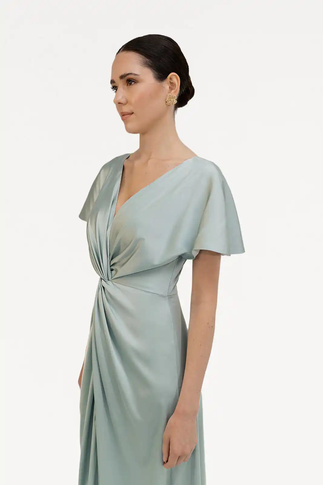 A woman with dark hair in a low bun wears the TH&TH Camilla Bridesmaid Dress in Dusty Eucalyptus Satin—this size-inclusive gown features short sleeves and a twist-knot bodice, shown against a plain white background.