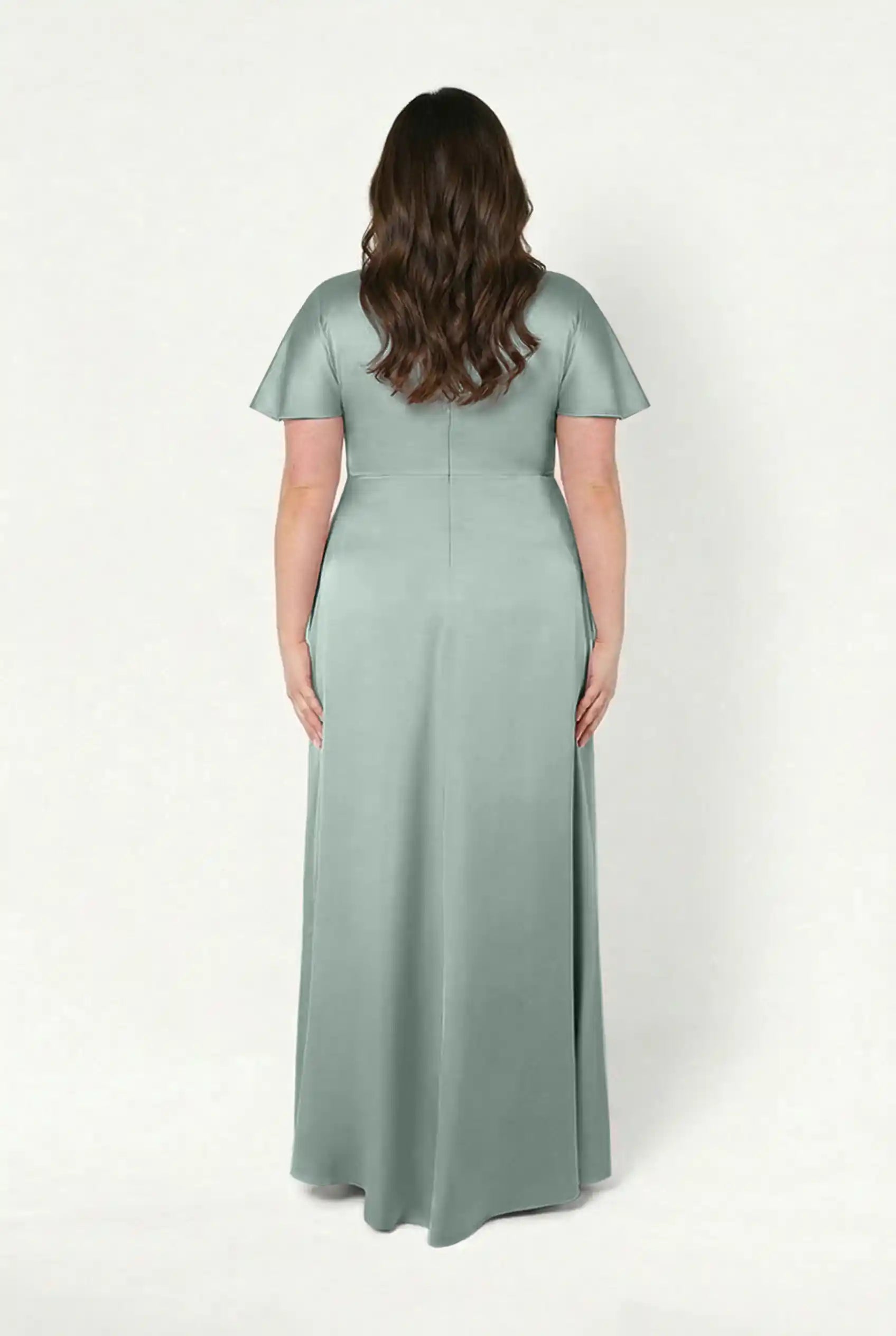 A woman with long, wavy brown hair stands facing away, wearing the TH&TH Camilla Bridesmaid Dress in floor-length, short-sleeved Dusty Eucalyptus satin with a twist-knot bodice against a white background.