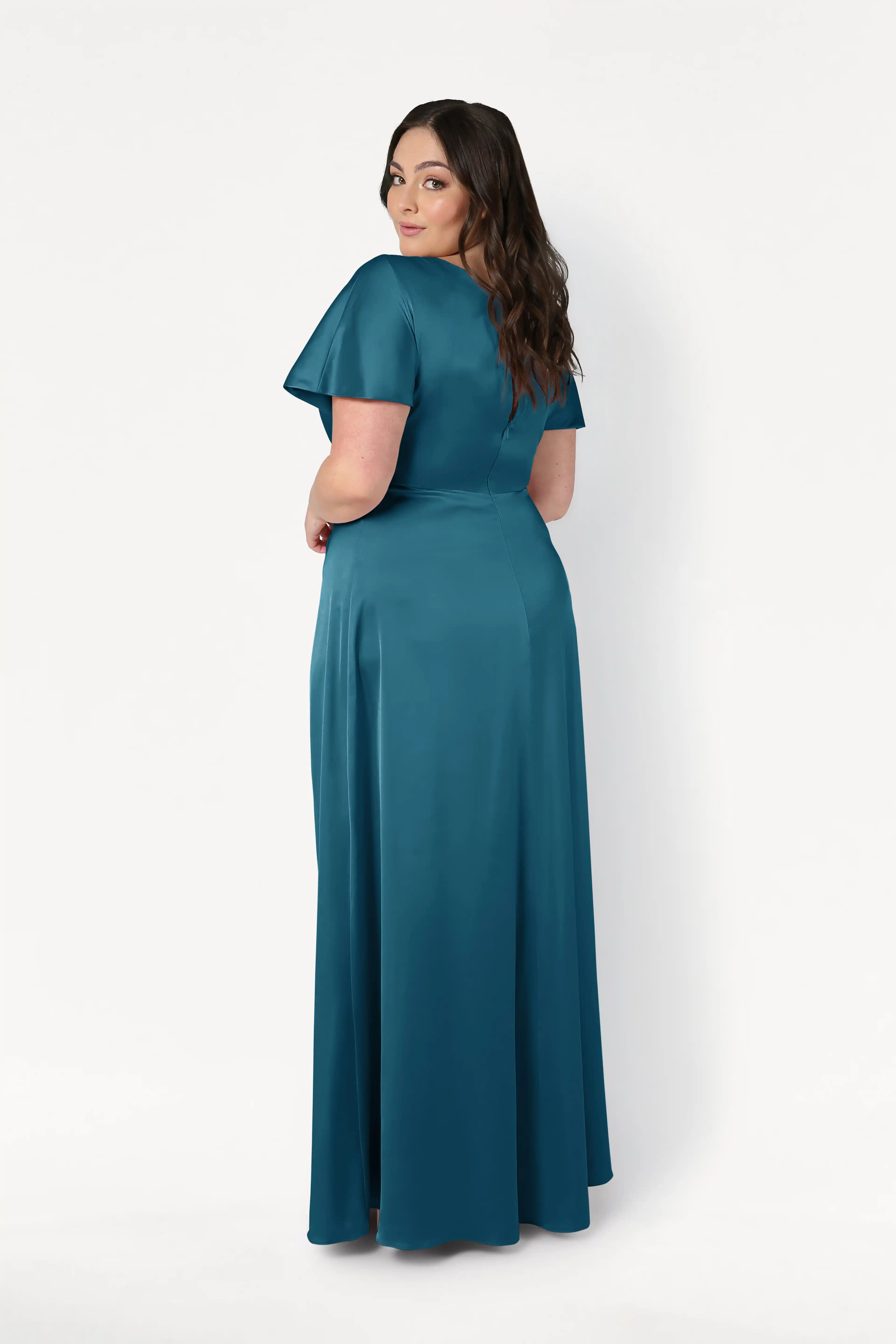 A woman with long dark hair stands, looking over her shoulder in the floor-length Camilla Bridesmaid Dress in Emerald Dawn Satin by TH&TH. The dress has short sleeves and is set against a plain white background.