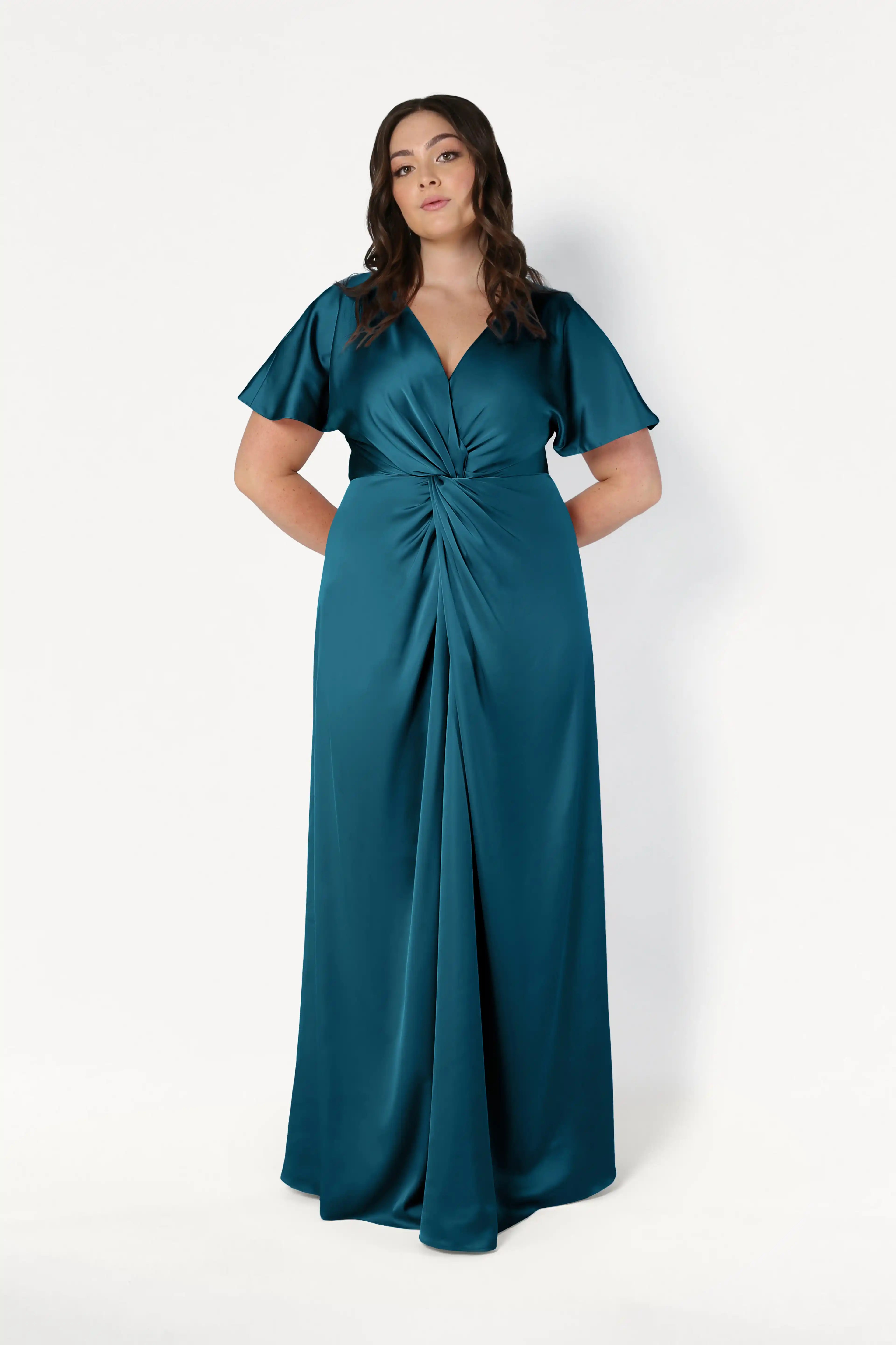 A woman with long wavy hair wears the TH&TH Camilla Bridesmaid Dress in Emerald Dawn Satin, featuring short flutter sleeves, a V-neckline, and a gathered twist waist, standing against a plain white background.