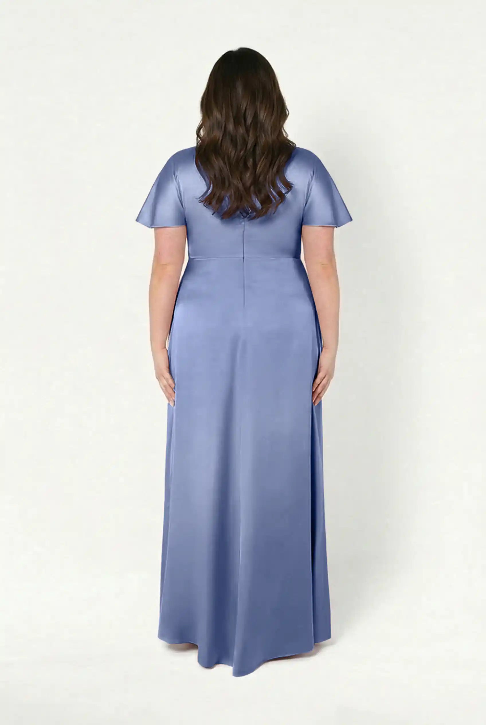 A woman with wavy brown hair is seen from behind wearing the TH&TH Camilla Bridesmaid Dress in French Blue Satin, a size-inclusive, floor-length gown with short sleeves, set against a plain light background.