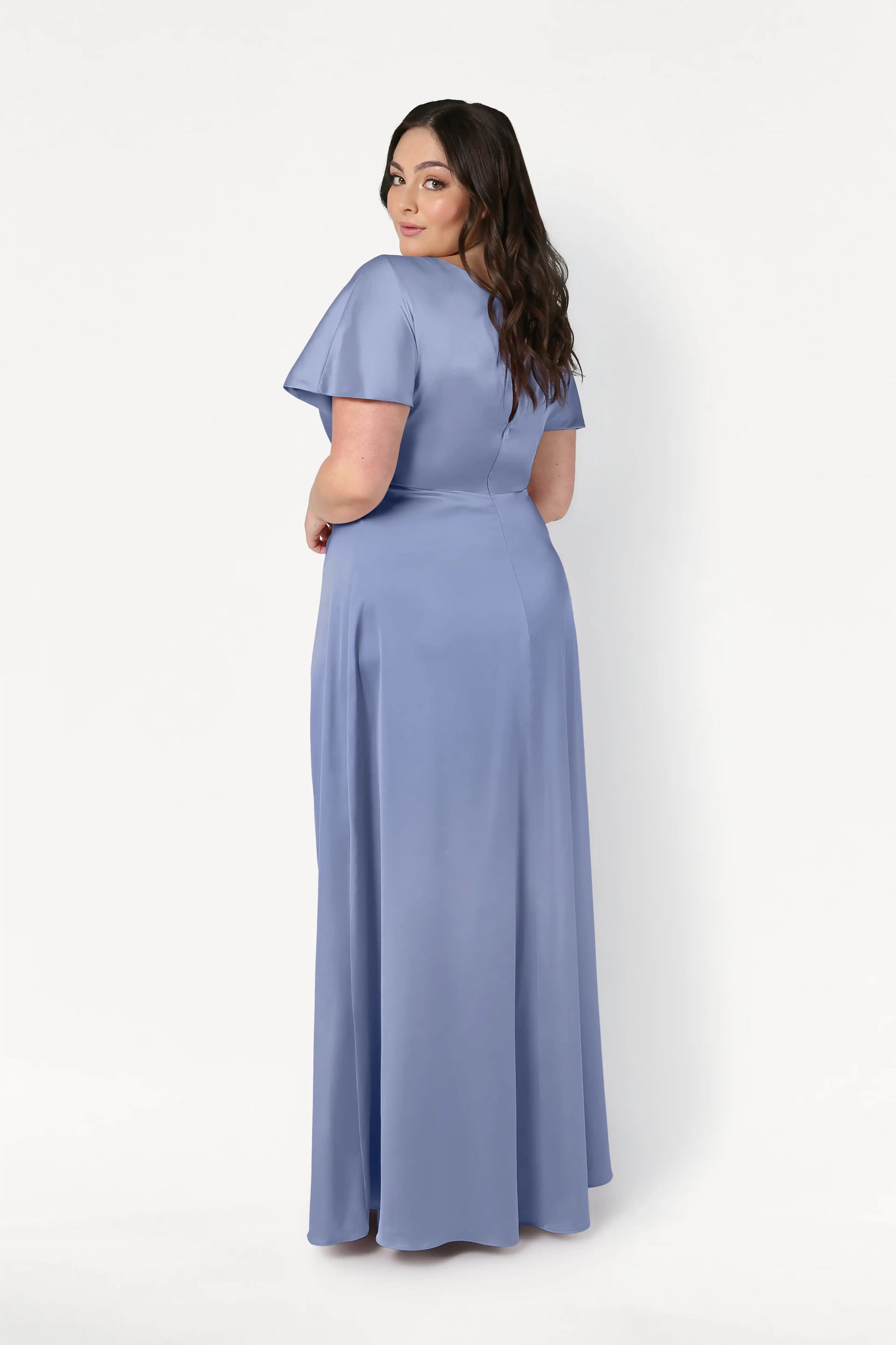 A woman with long dark hair stands facing away, looking over her shoulder in the TH&TH Camilla Bridesmaid Dress in French Blue Satin—a floor-length, short-sleeve gown with a twist-knot bodice—set against a plain white background.