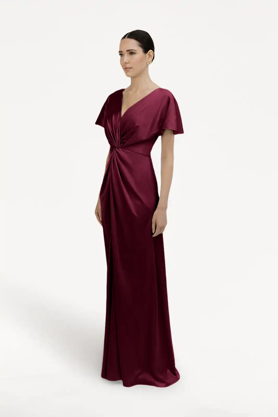 A woman wears the TH&TH Camilla Bridesmaid Dress in Merlot Satin, a floor-length modern gown with short sleeves, V-neckline, and twisted knot waist; her dark hair is pulled back as she faces slightly sideways against a plain white background.
